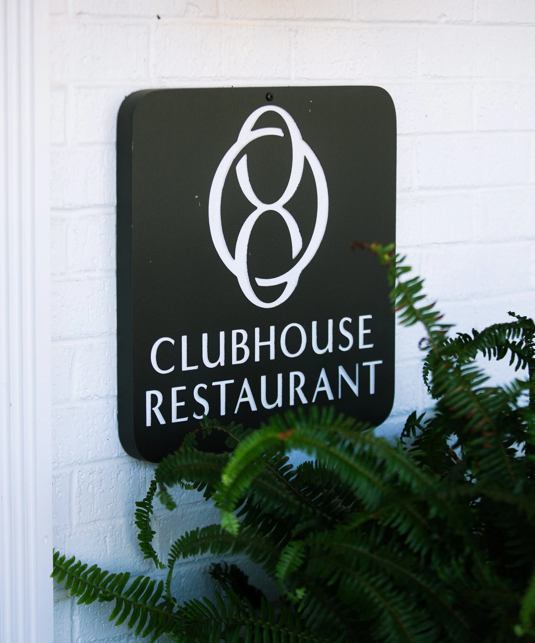 Sign on white brick wall with black background and white logo for clubhouse, restaurant, and festival, partially obscured by green ferns