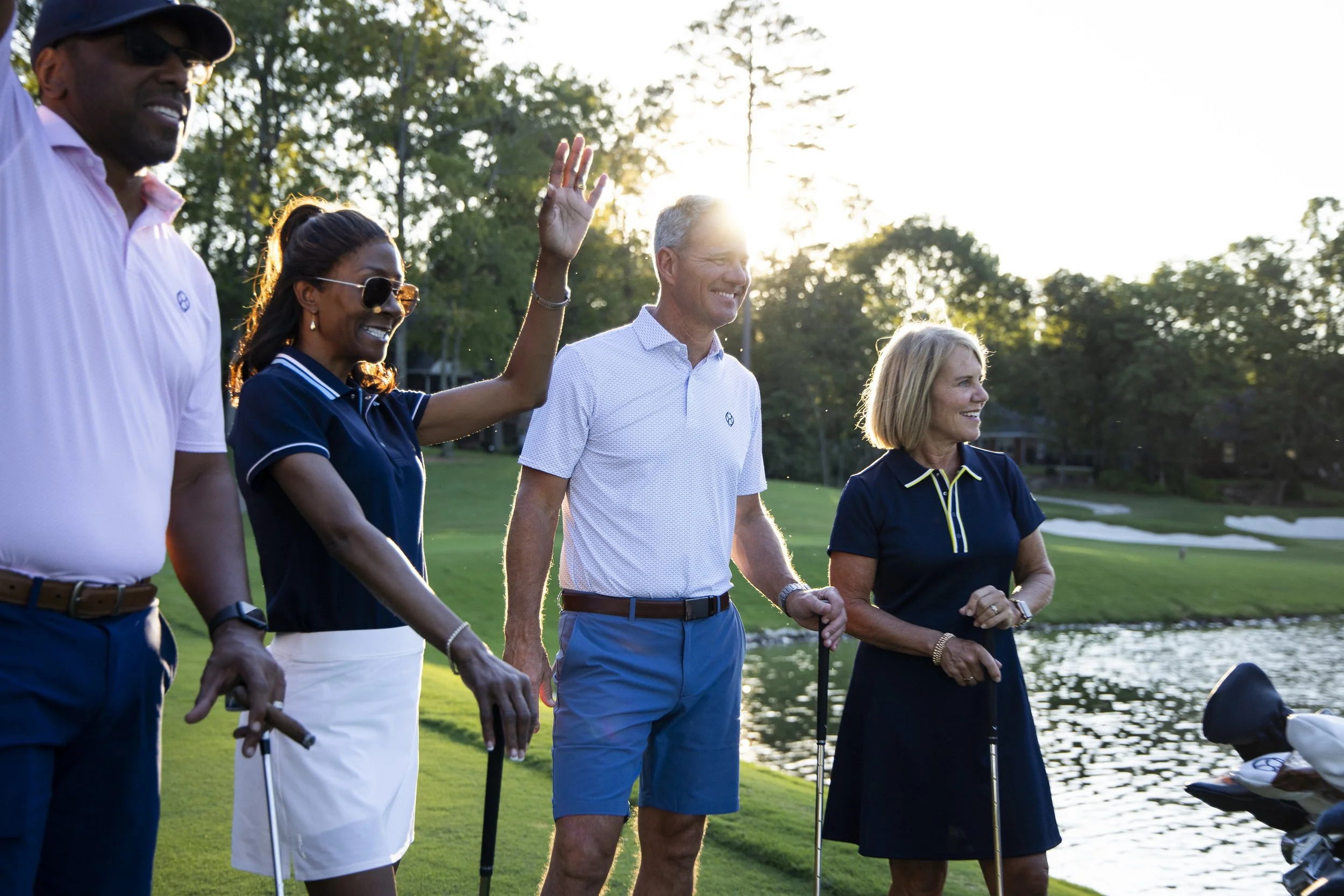Group of five people walking near a pond on a golf course during sunset, dressed in casual golf attire, smiling and holding golf clubs.