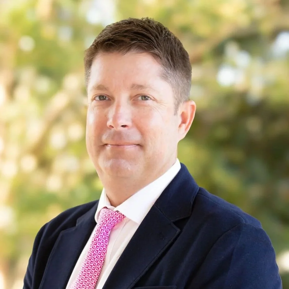 A professional man outdoors with a blurred green and yellow background, wearing a dark blazer, white shirt, and pink tie.