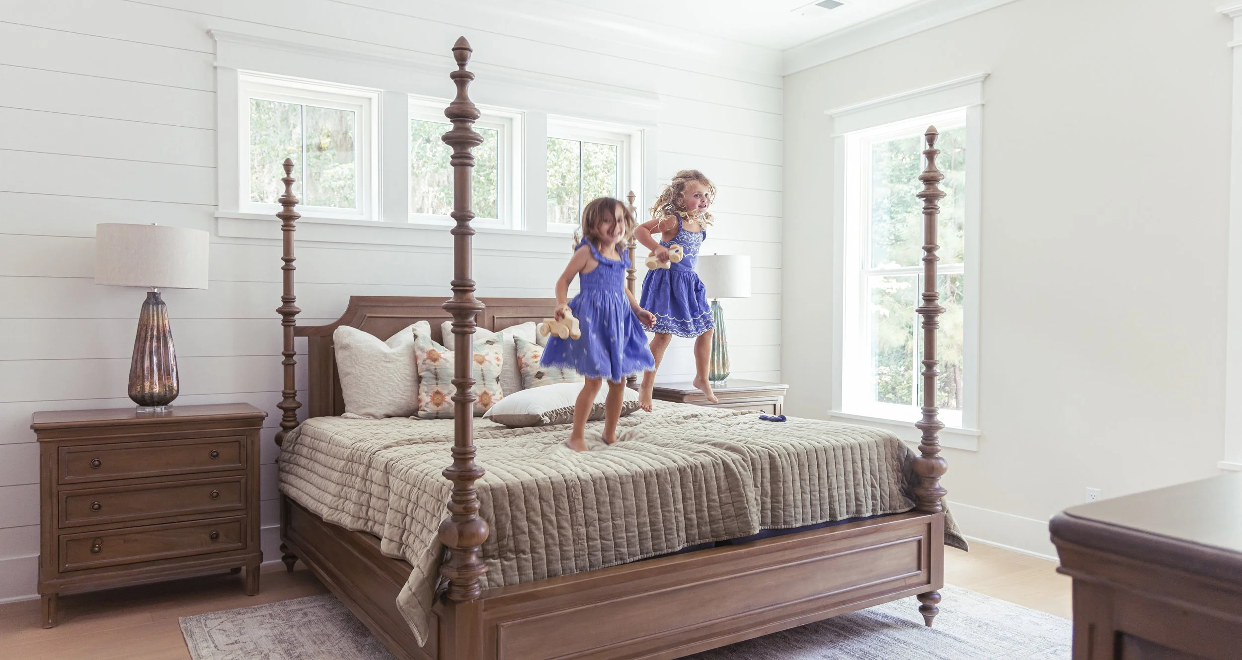 Two young girls in blue dresses jumping on a bed in bright bedroom with white walls and large windows, wooden furniture, and decorative pillows.