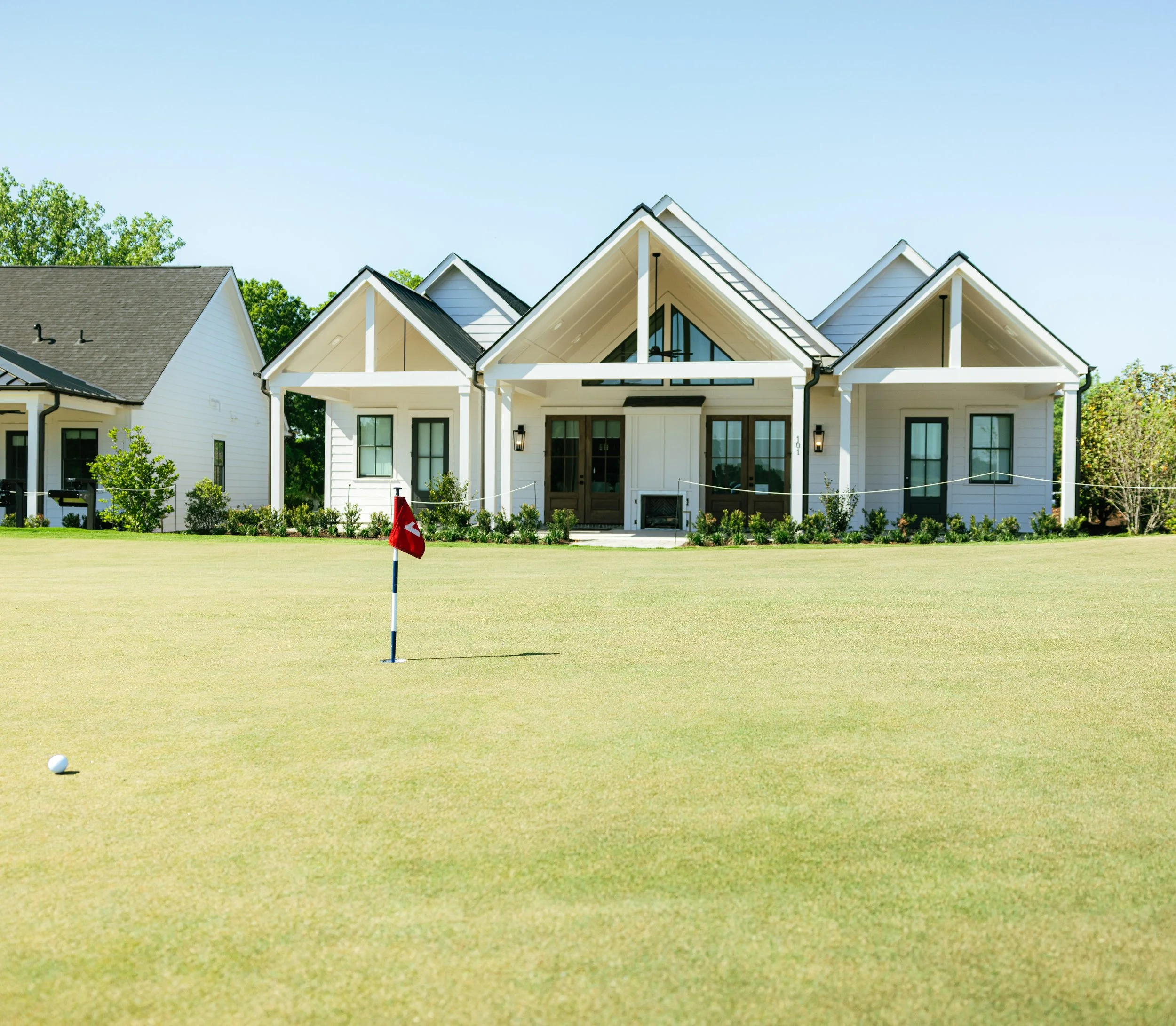 A golf course with a putting green, a white golf ball, and a flagstick with a red flag, with a white house and trees in the background.