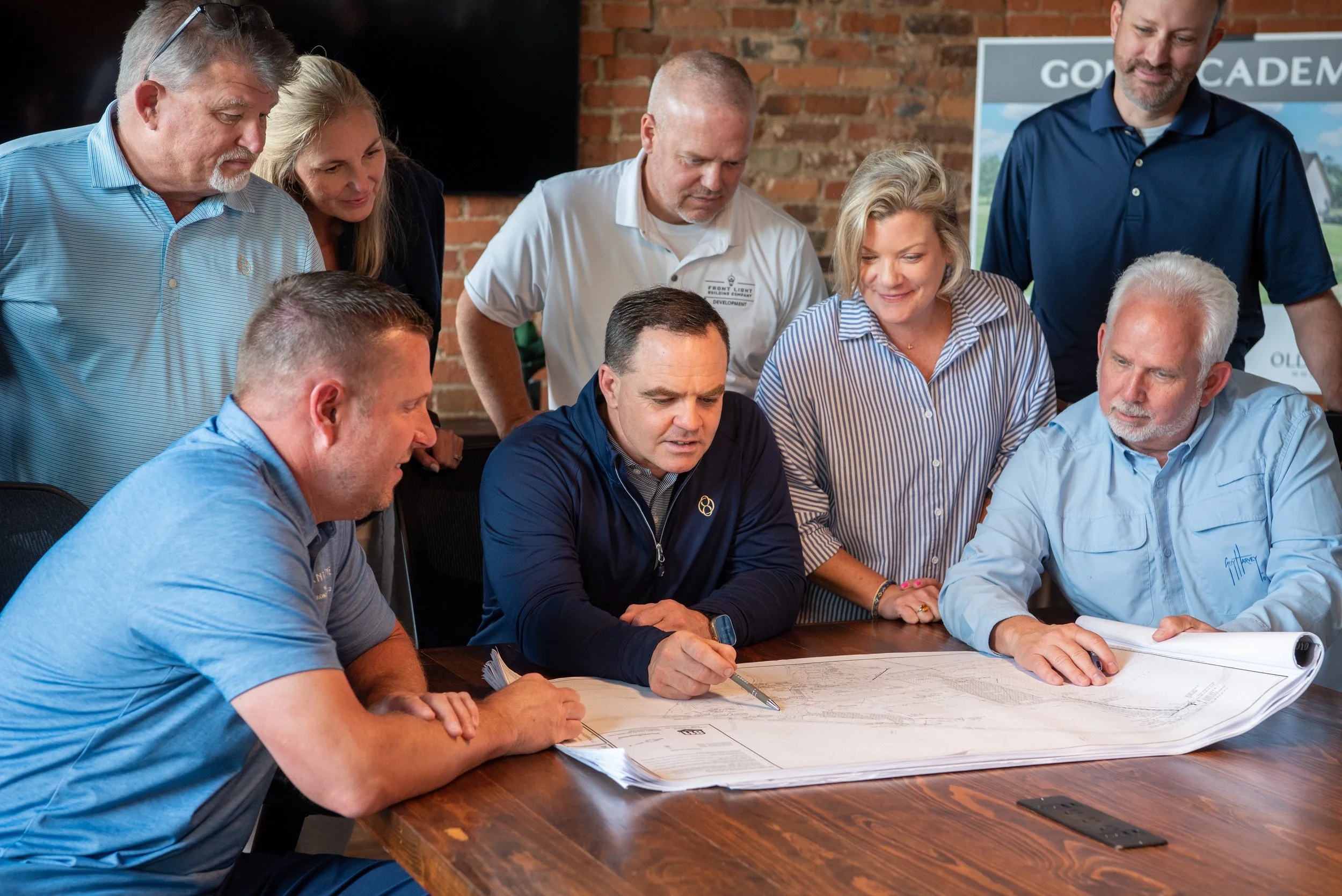 A group of eight people gathered around a table, looking at documents. They appear to be in a meeting, with some standing and some sitting, in a room with a brick wall background.