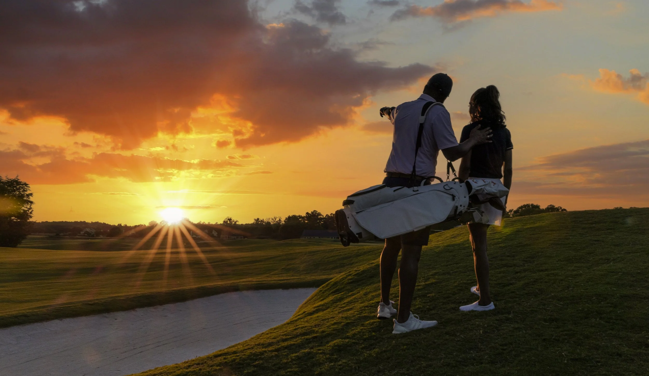 A man and woman standing on a golf course during sunset, with the man pointing towards the horizon as the woman looks on, both carrying golf bags.