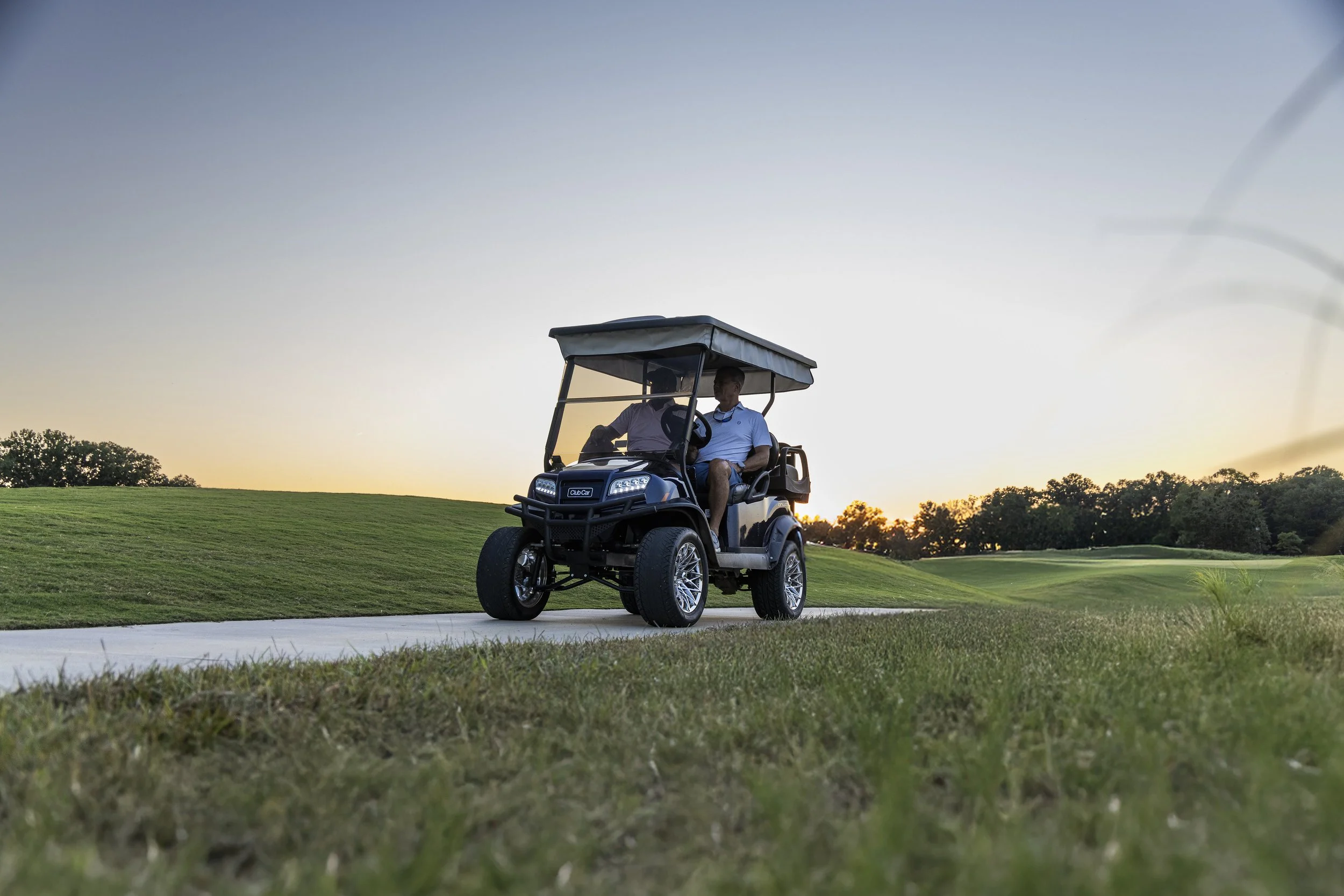 Two men riding in a golf cart on a golf course during sunset.