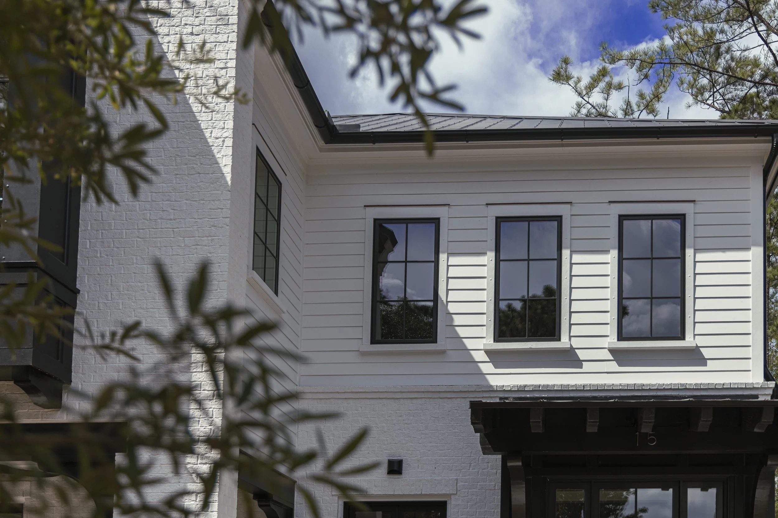 Part of a white two-story house with black window frames, a section of white brick wall, and a section of white vertical siding. There are trees and a blue sky with some clouds in the background.