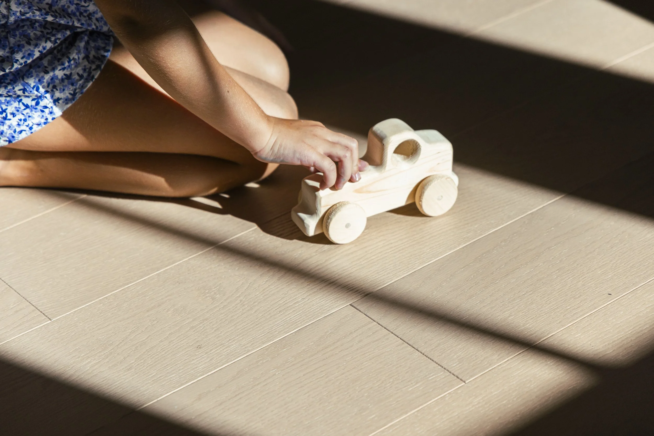 A person wearing a blue floral dress playing with a wooden toy train on a light-colored hardwood floor.