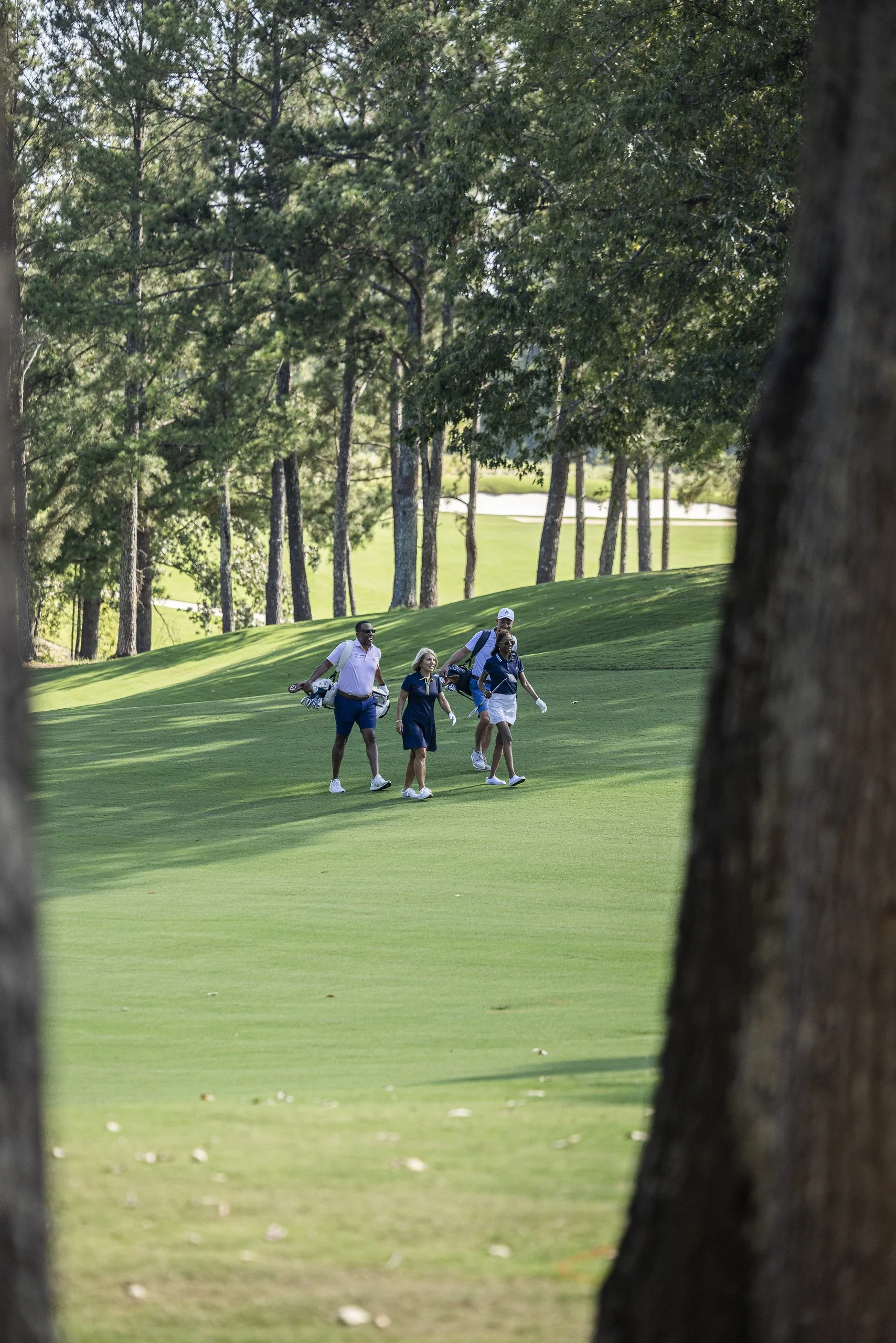 Four people walking on a golf course path through a wooded area, carrying golf clubs.