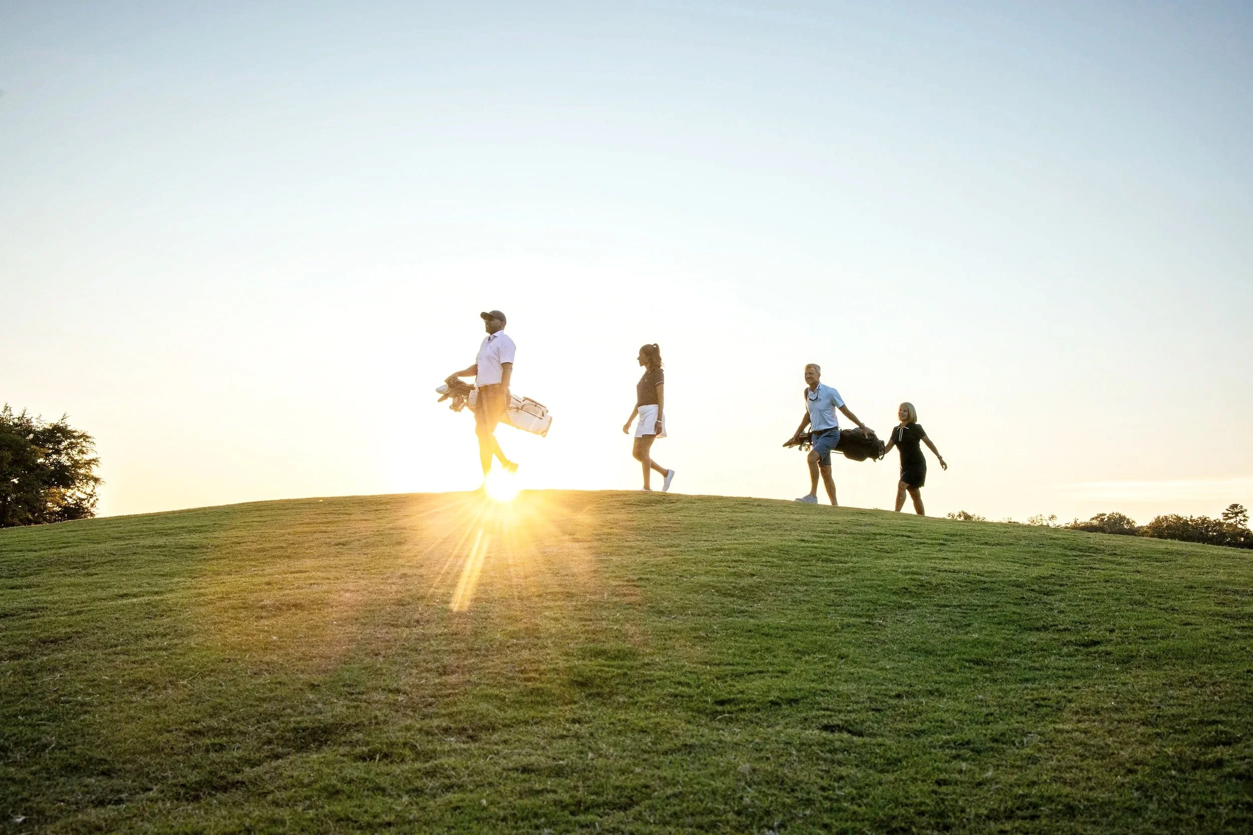 Four people walking uphill on a golf course at sunset, carrying golf clubs and bags.