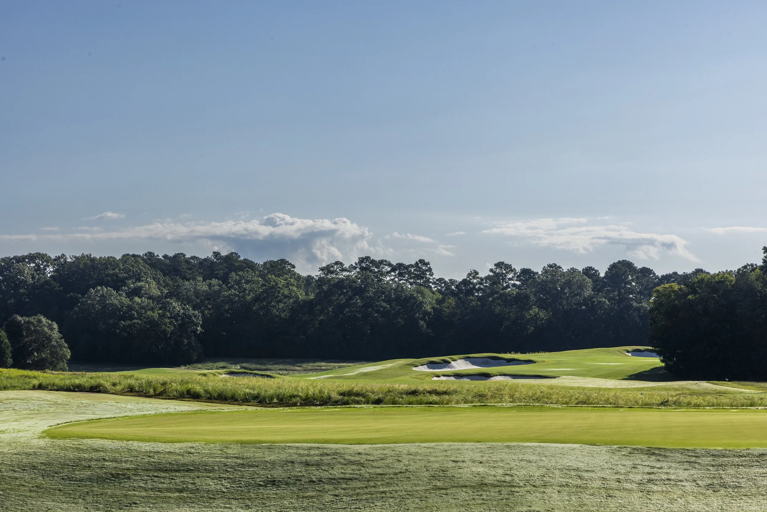 A golf course with green fairways, sand bunkers, and a dense treeline in the background under a partly cloudy sky.