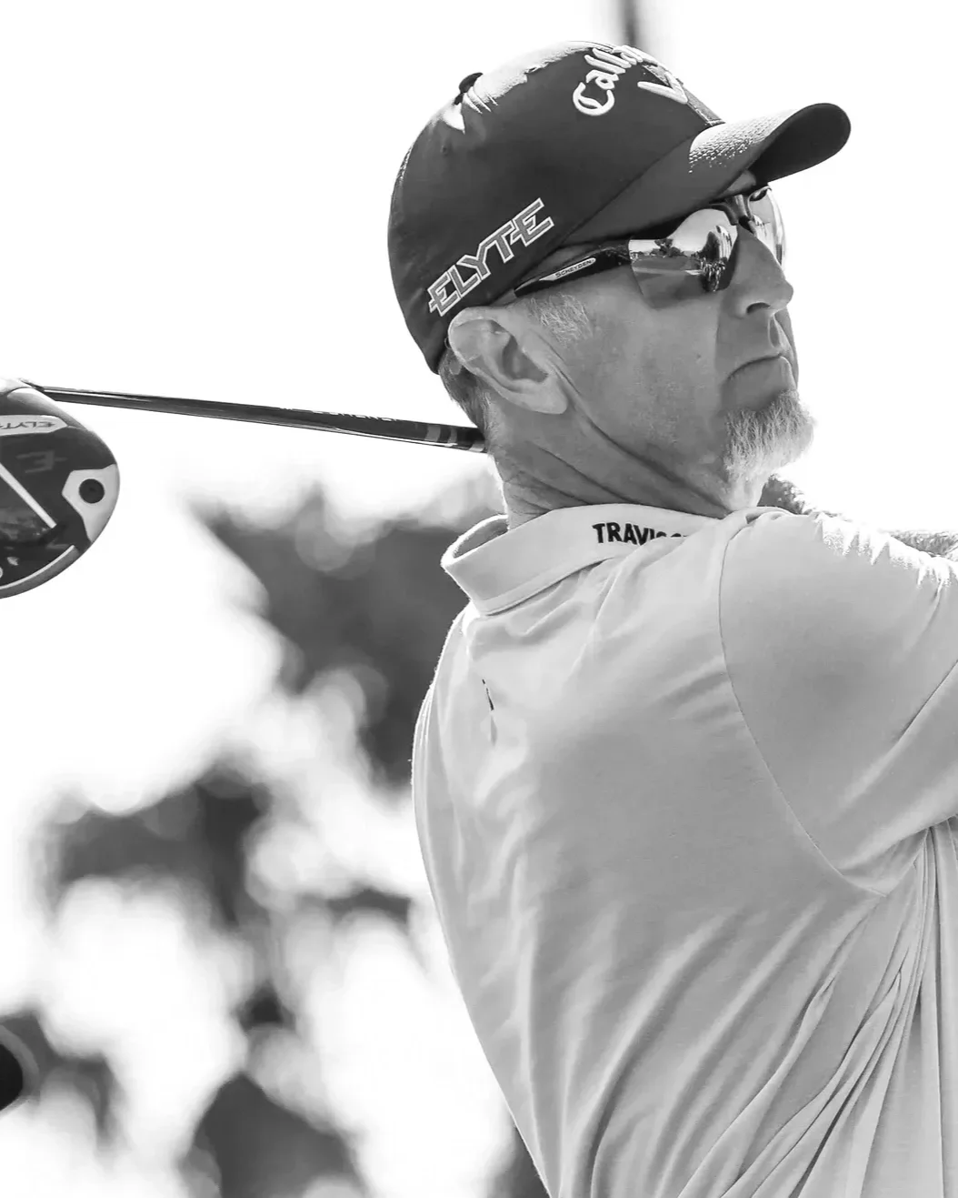 A male golfer wearing a Callaway cap, sunglasses, and a light-colored polo shirt, swings a golf club on a golf course.