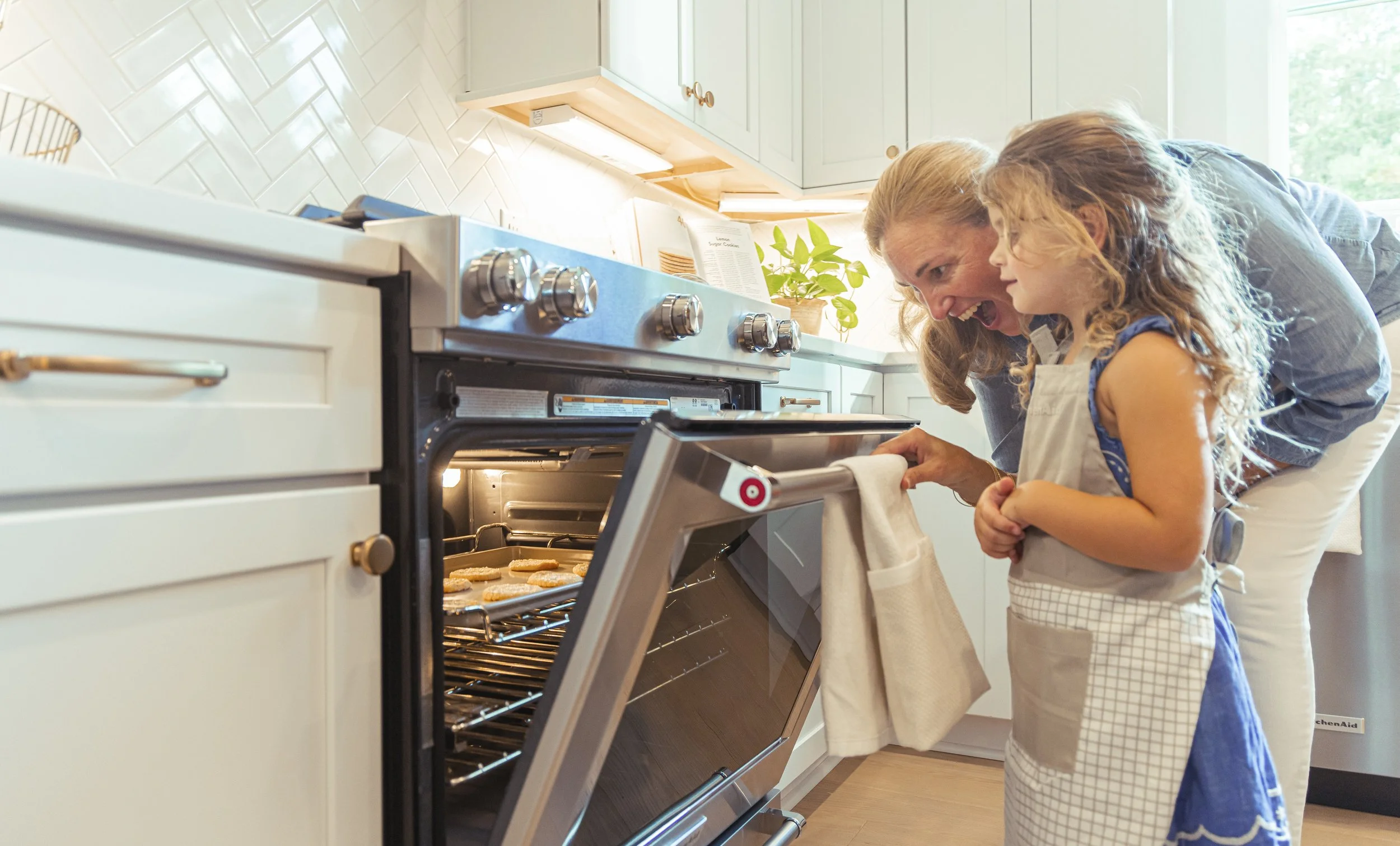A woman and a young girl baking cookies together in a cozy kitchen. The woman is leaning over the oven, smiling and talking to the girl who is holding a towel. The oven door is open, revealing cookies inside.
