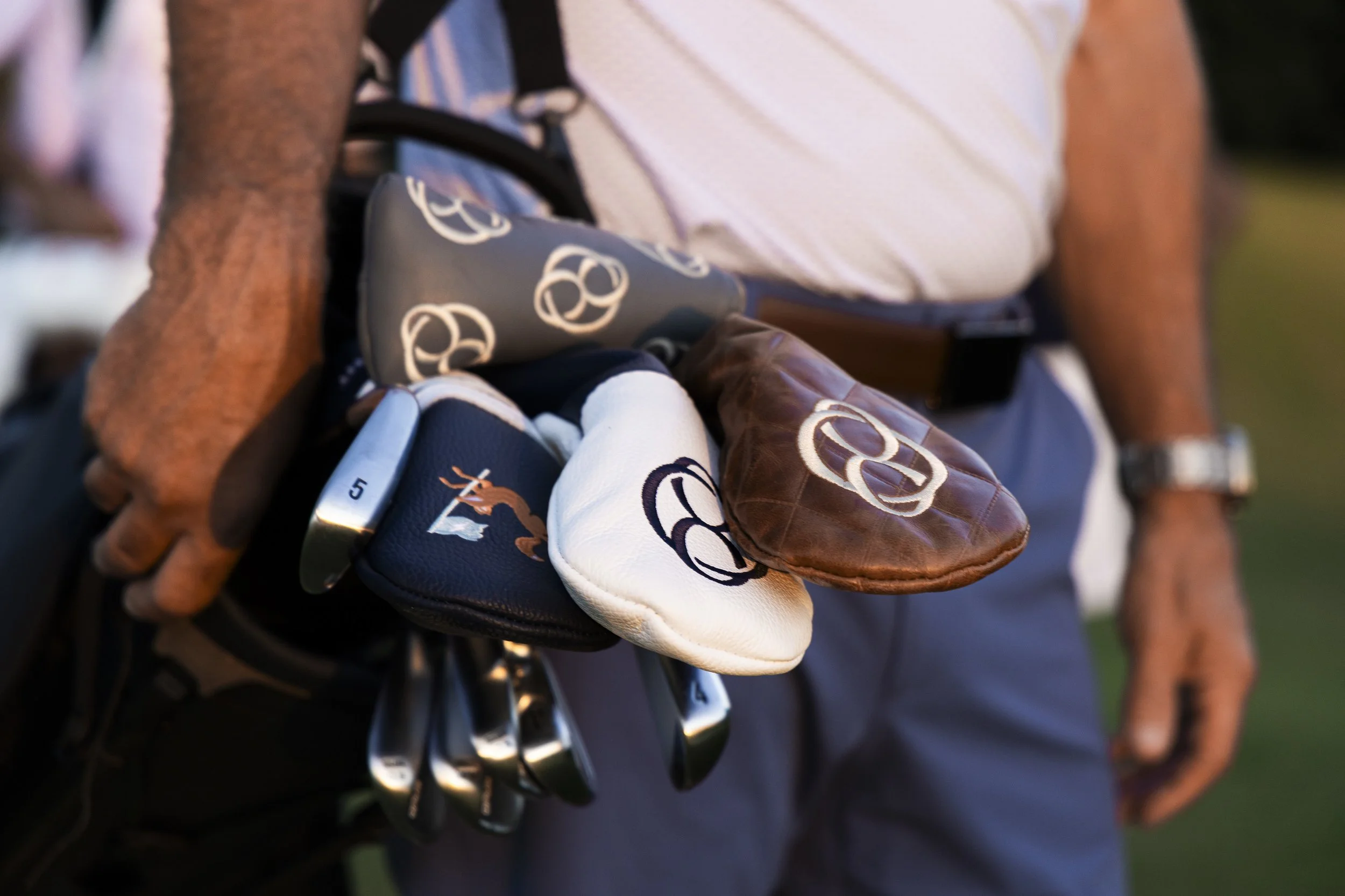 A person holding a set of golf clubs with golf gloves, with a focus on the clubheads and gloves featuring circular logos, outdoors during sunset.