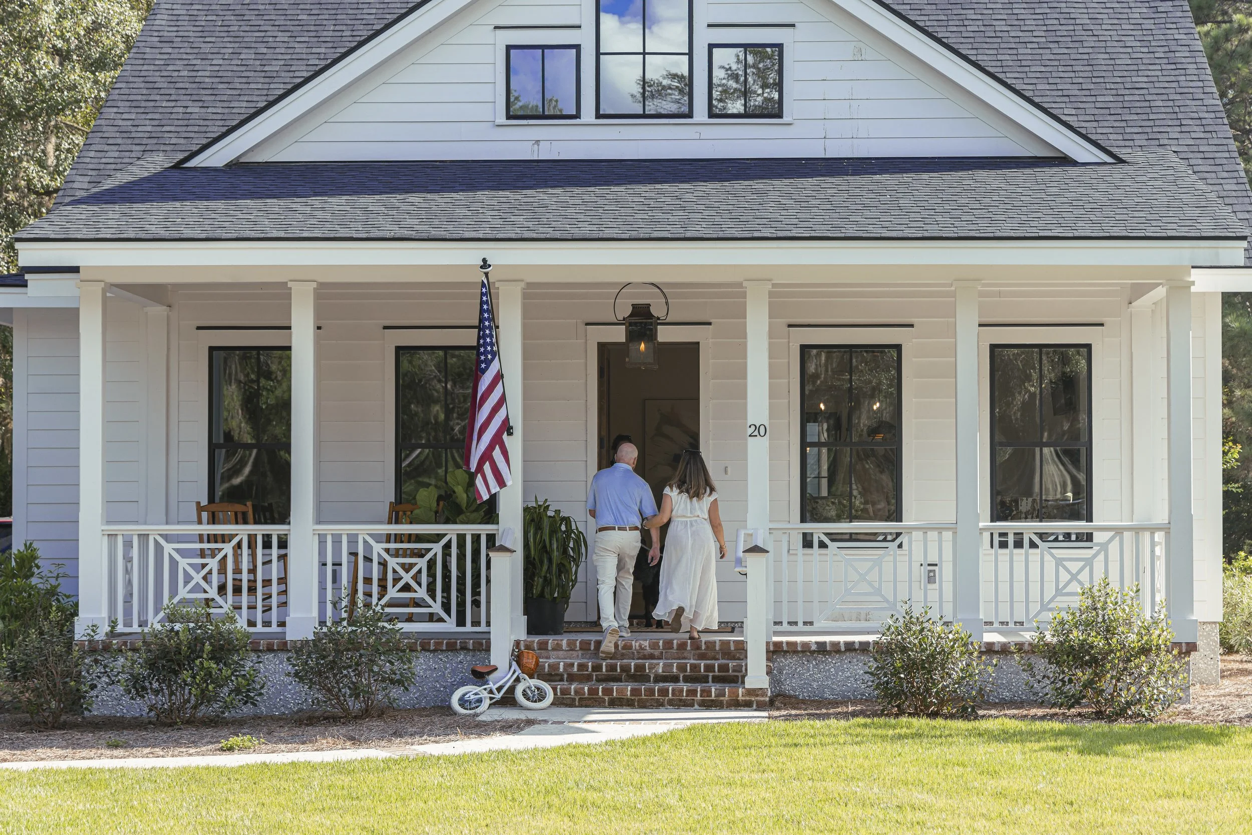 A white house with a front porch, two people walking up the steps towards the entrance, an American flag hanging, a small child's bicycle on the walk, and greenery in the yard.