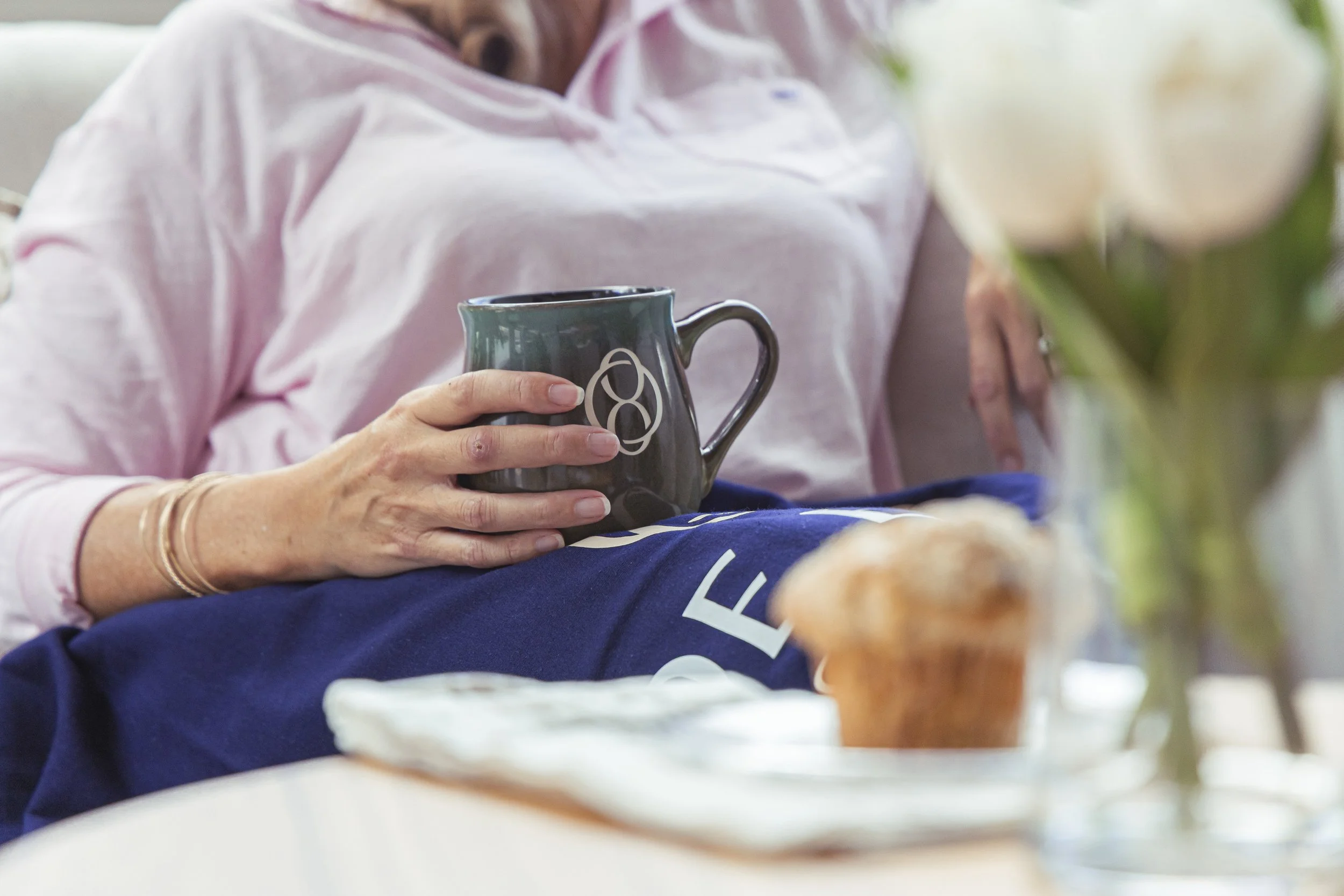 Close-up of a person holding a dark-colored mug with a white logo, sitting at a table with food, wearing a pink shirt, with a blurred background.