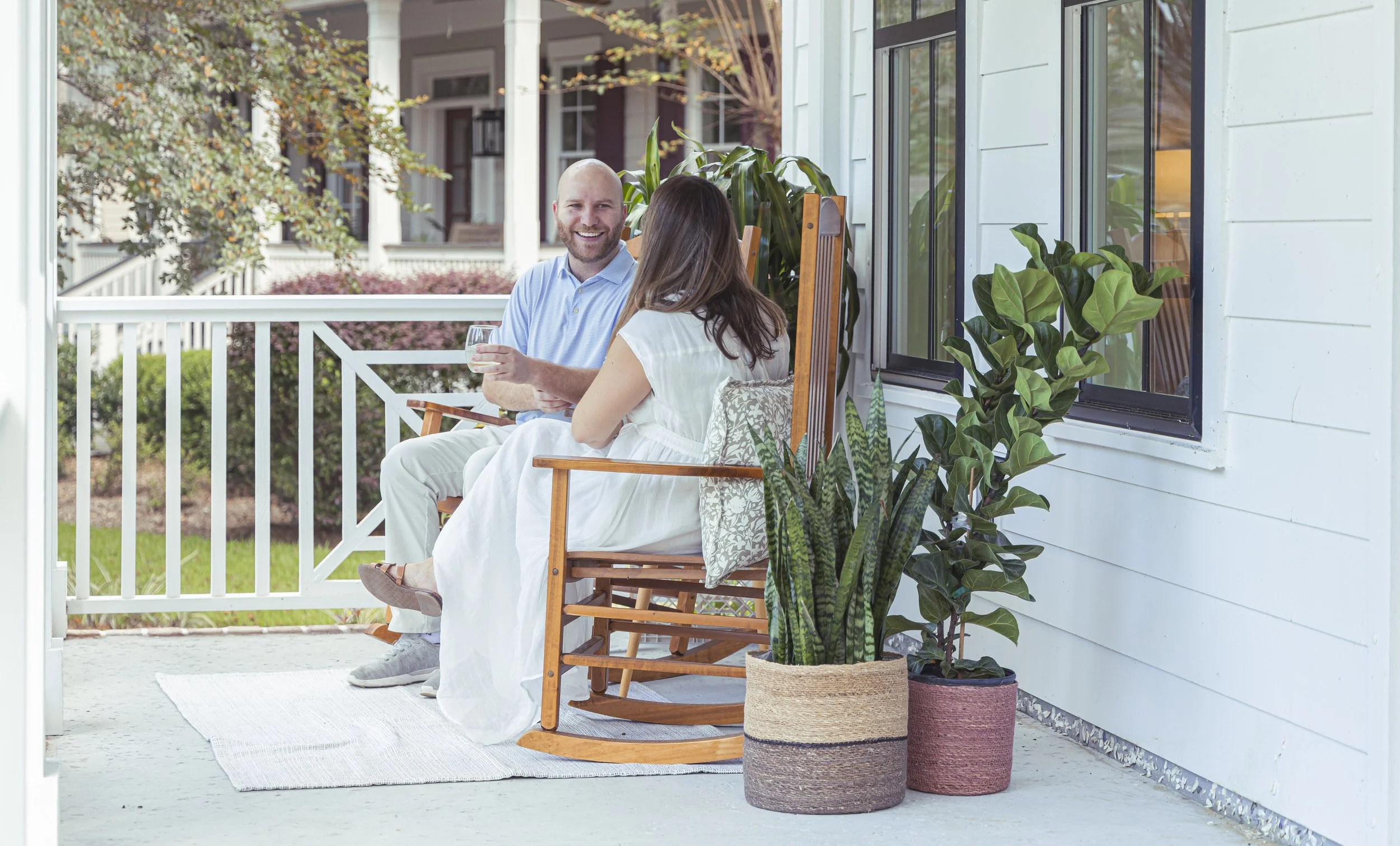 A man and a woman sitting on a porch swing, holding hands and smiling at each other, with houses and trees in the background and potted plants nearby.
