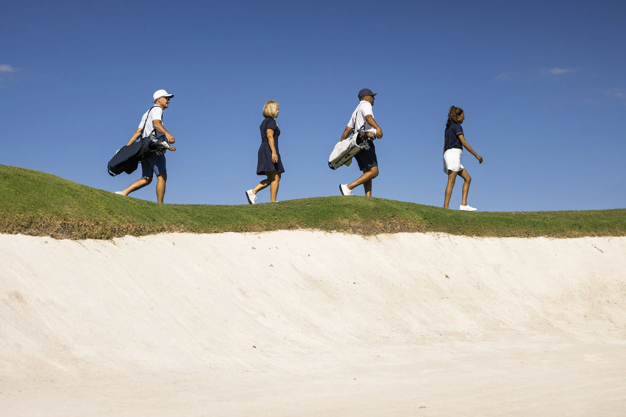 Four people walking on a grassy hill with golf clubs, blue sky in the background.