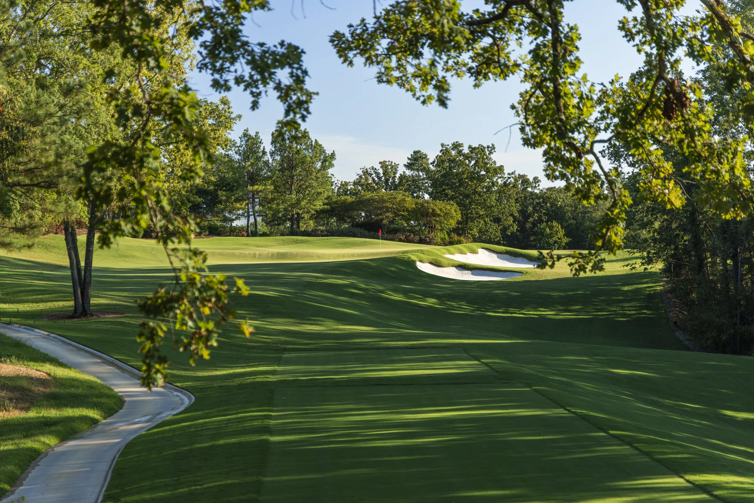 A scenic golf course with green fairways, sand traps, and a red flag, framed by trees under a clear blue sky.