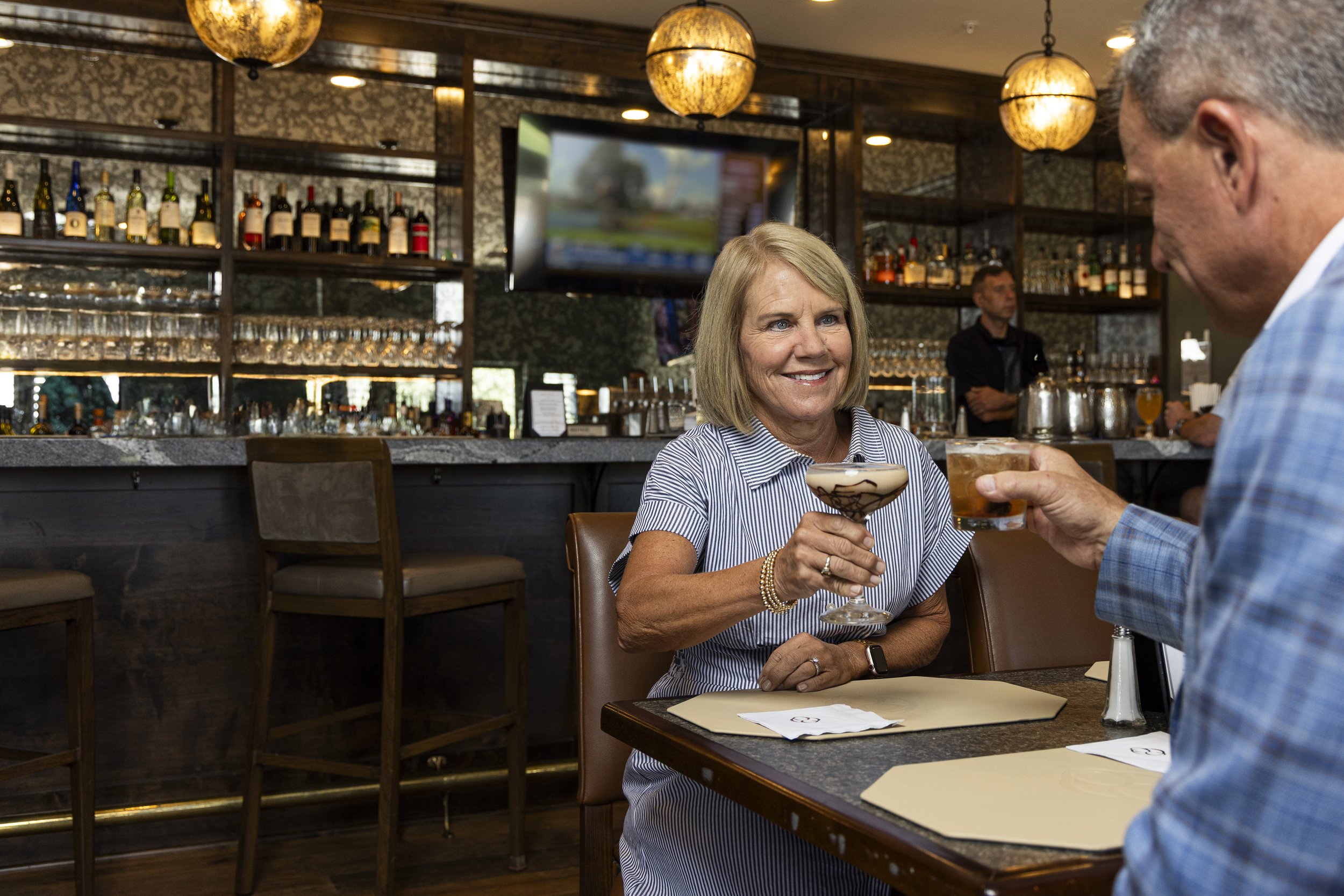 A woman with blonde hair holding a cocktail glass, smiling, across from an older man at a restaurant table.