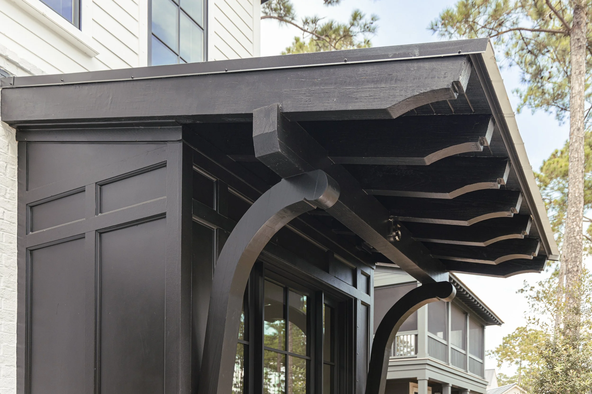 Close-up of a black wooden pergola with curved beams in front of a modern house with large windows and a white exterior, surrounded by trees.
