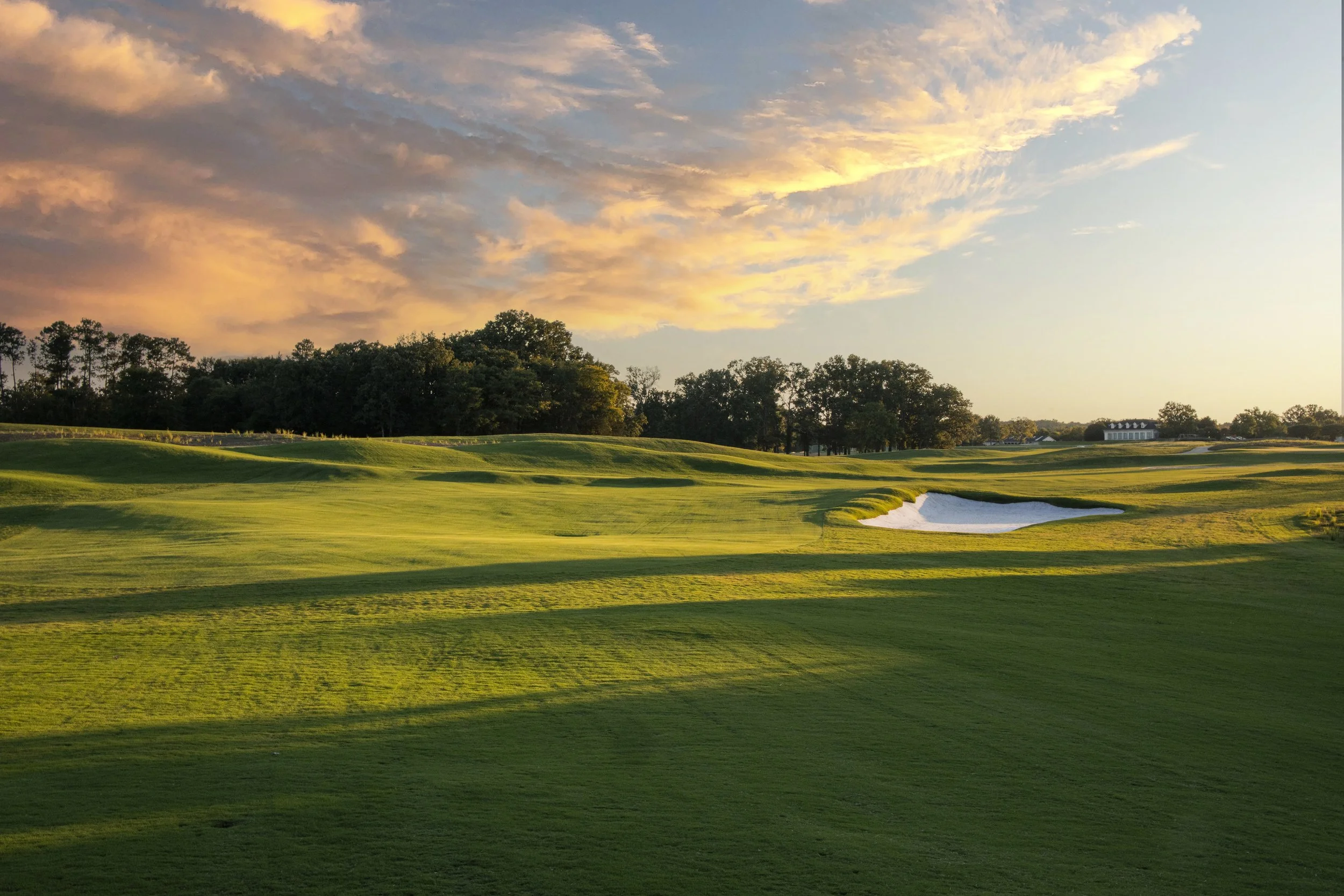 Sunset over a golf course with green fairways, trees, sand traps, and a clubhouse in the distance.