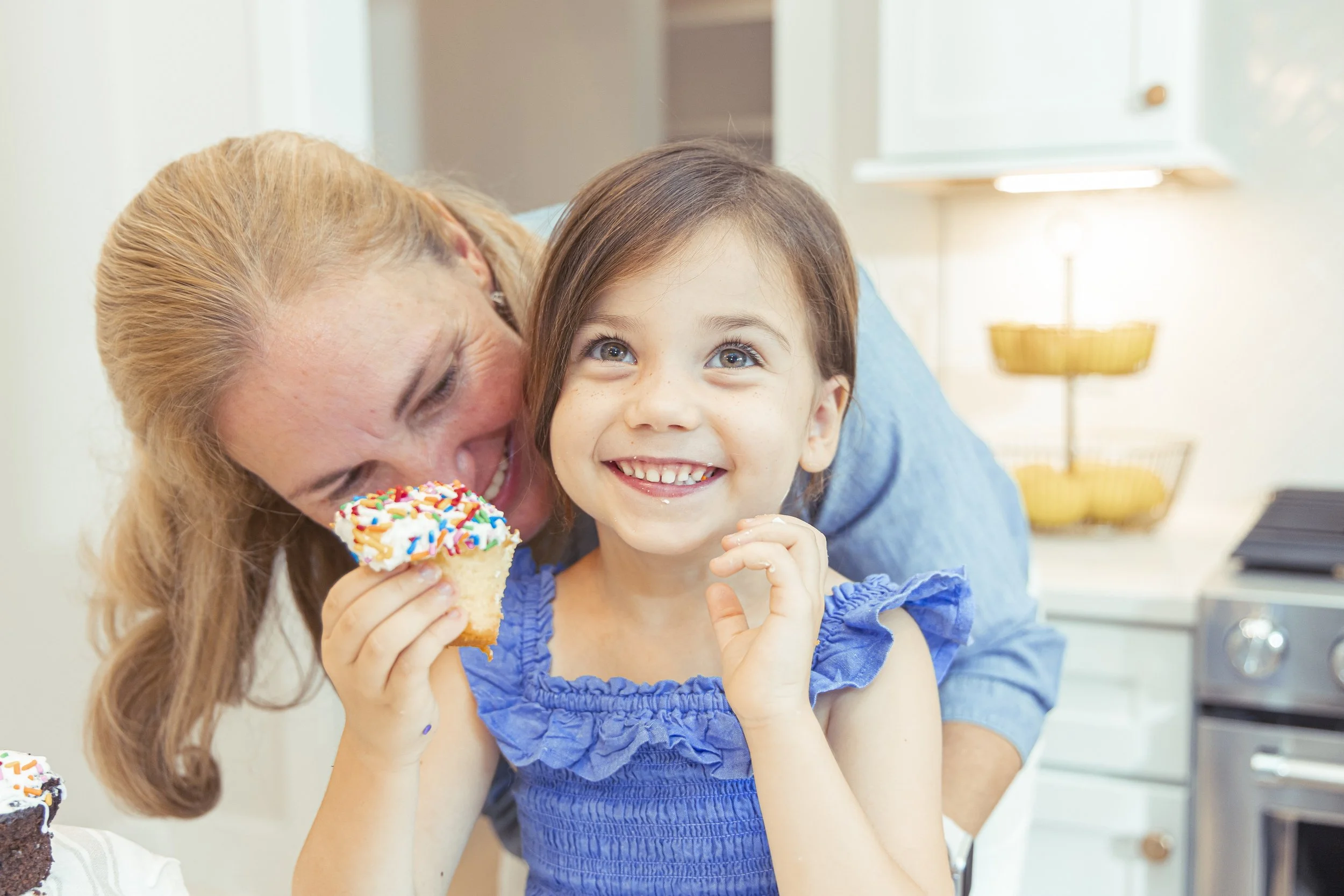 A young girl with brown hair and blue eyes smiling while holding a cupcake with colorful sprinkles, accompanied by an older woman with blonde hair who is also smiling, in a well-lit kitchen.