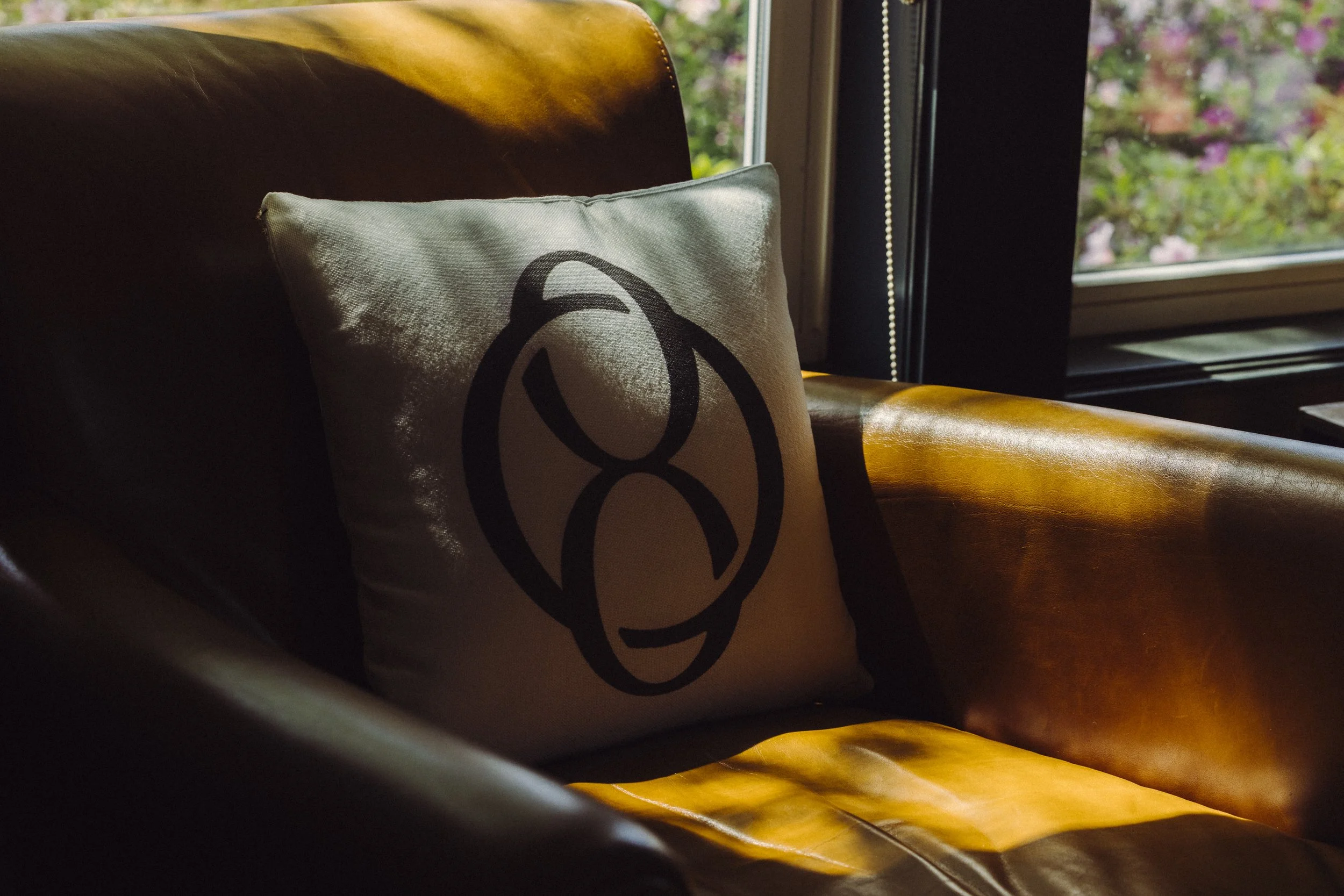 A decorative pillow with a black abstract design on a white or light-colored fabric, resting on a brown leather sofa next to a window with a view of colorful flowers outside.