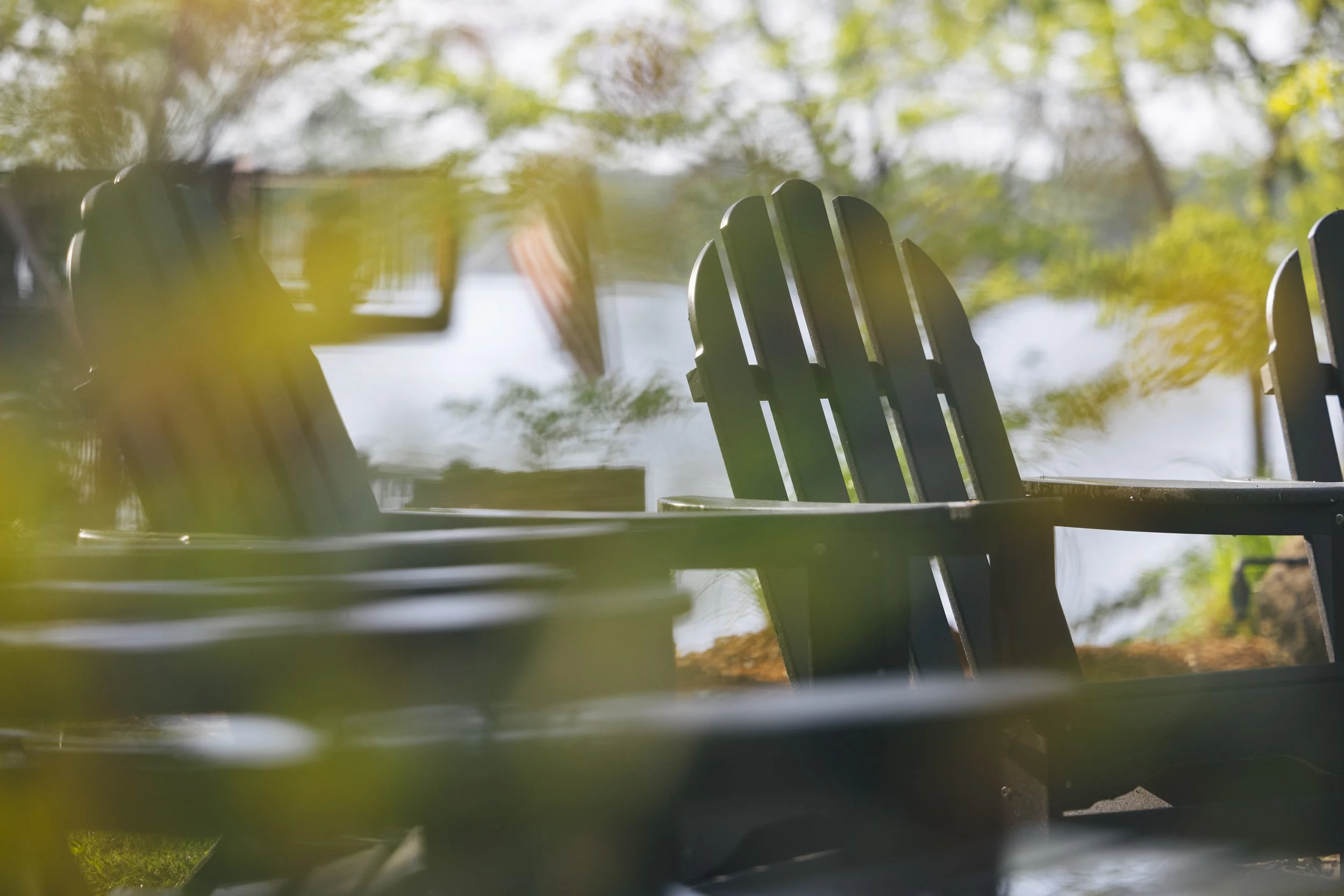 Empty black Adirondack chairs outdoors near a lake or pond with trees and sunlight in the background.