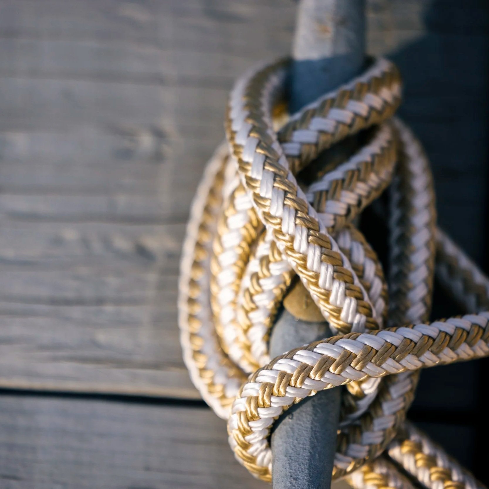 A rope ladder made of white and tan braided material hanging on a metal pole, with a weathered wooden wall in the background.
