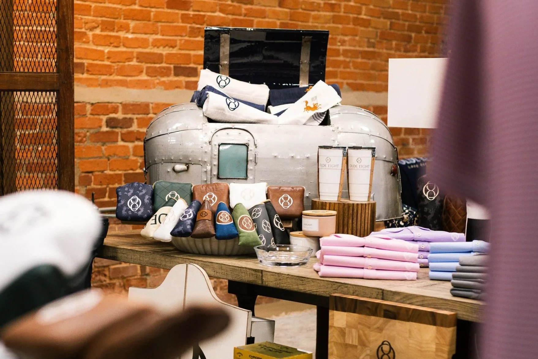 Display of various branded merchandise including hats, cups, pouches, and folded shirts or textiles on a wooden table against a brick wall.