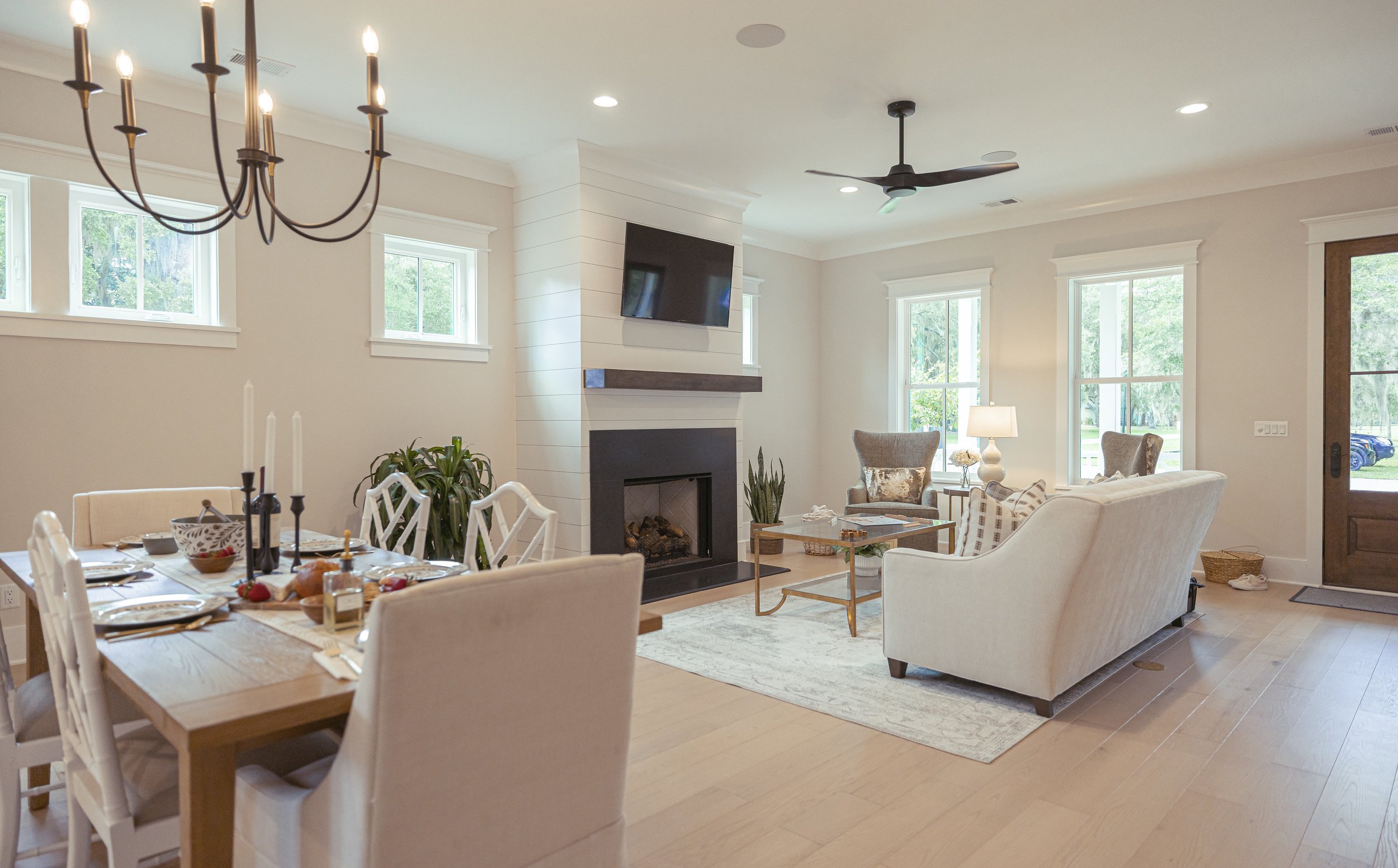 Living room with a dining table set for a meal, fireplace, television, ceiling fan, large windows, and comfortable chairs.