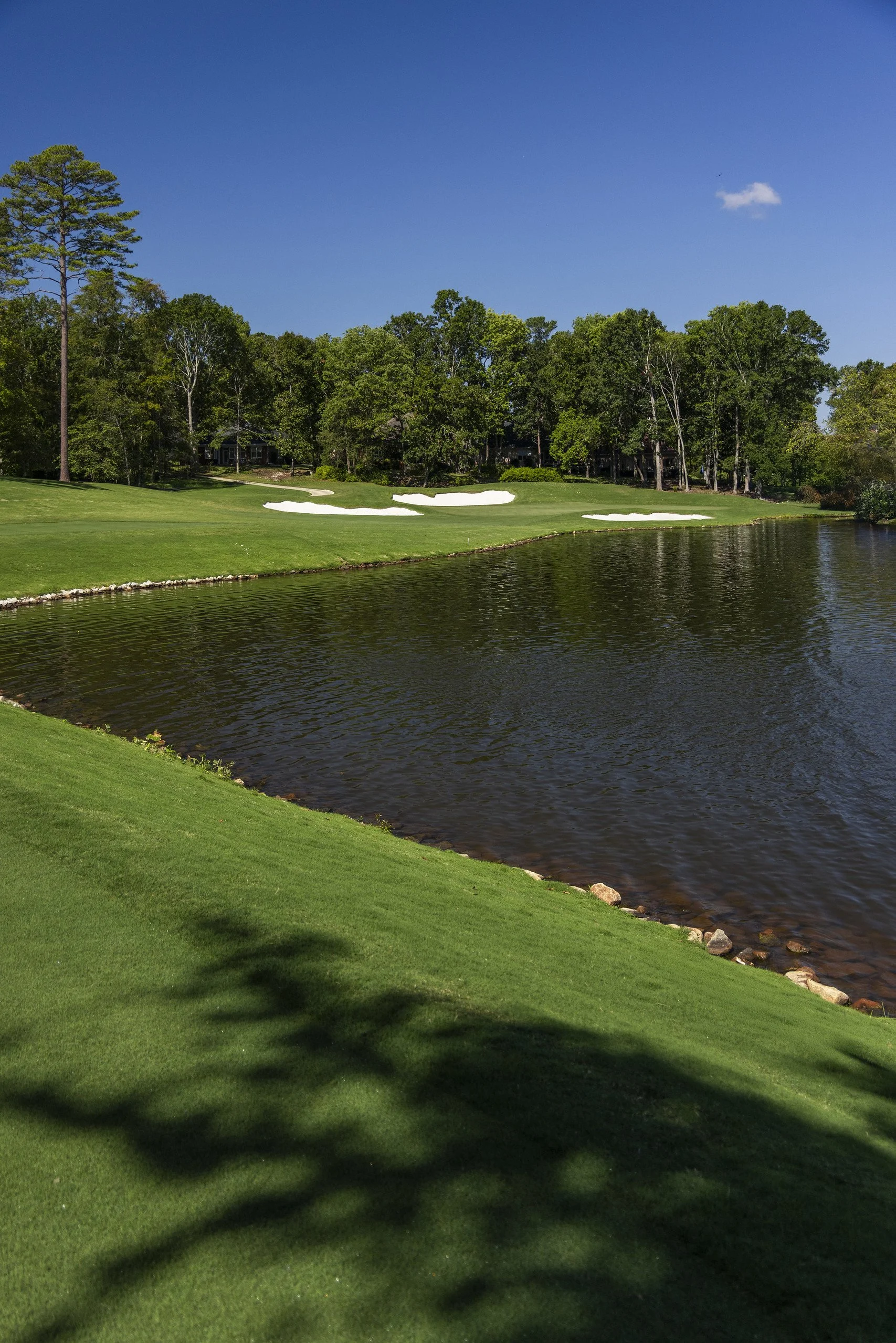 A golf course with a green fairway, sand bunkers, a water hazard, and trees in the background under a blue sky.