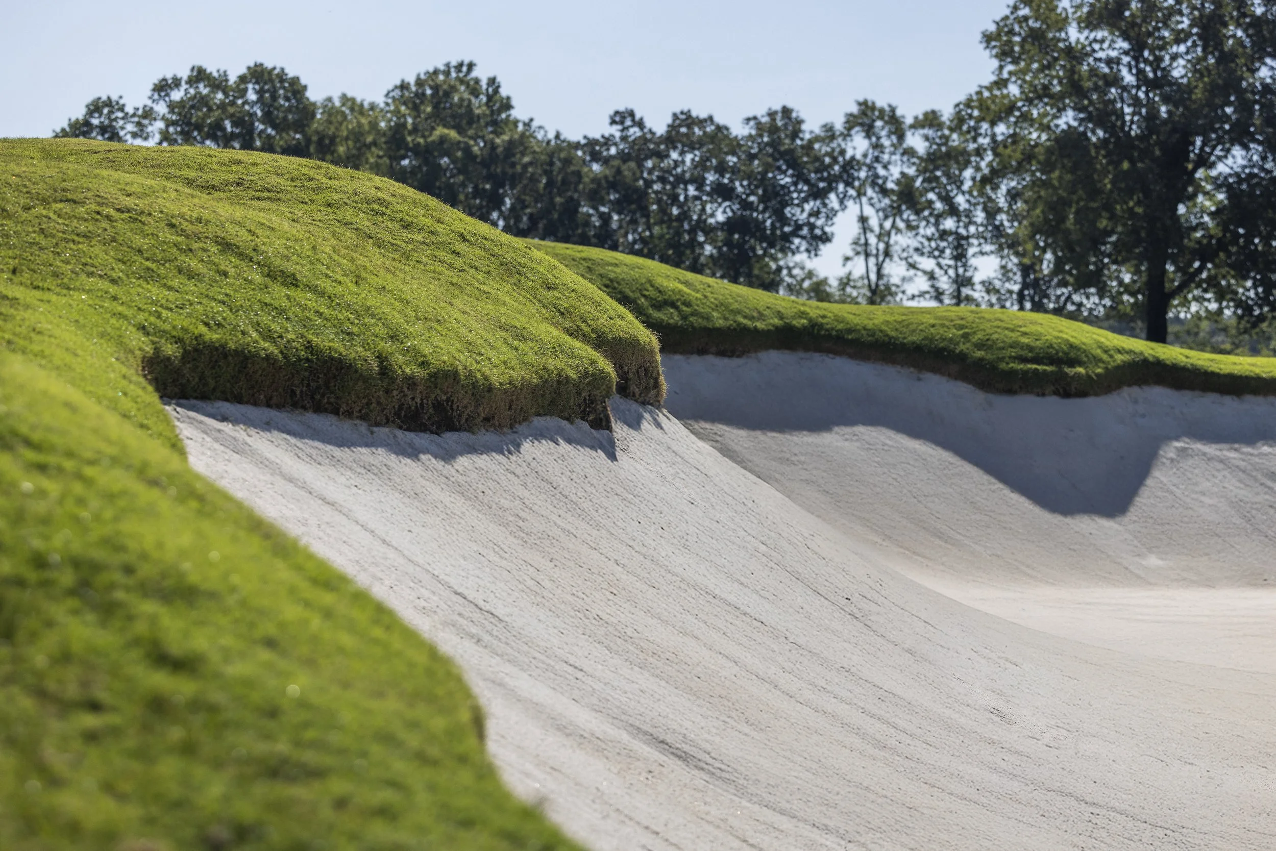 Close-up view of a golf bunker with smooth, white sand and well-maintained green grass around it, with trees in the background.
