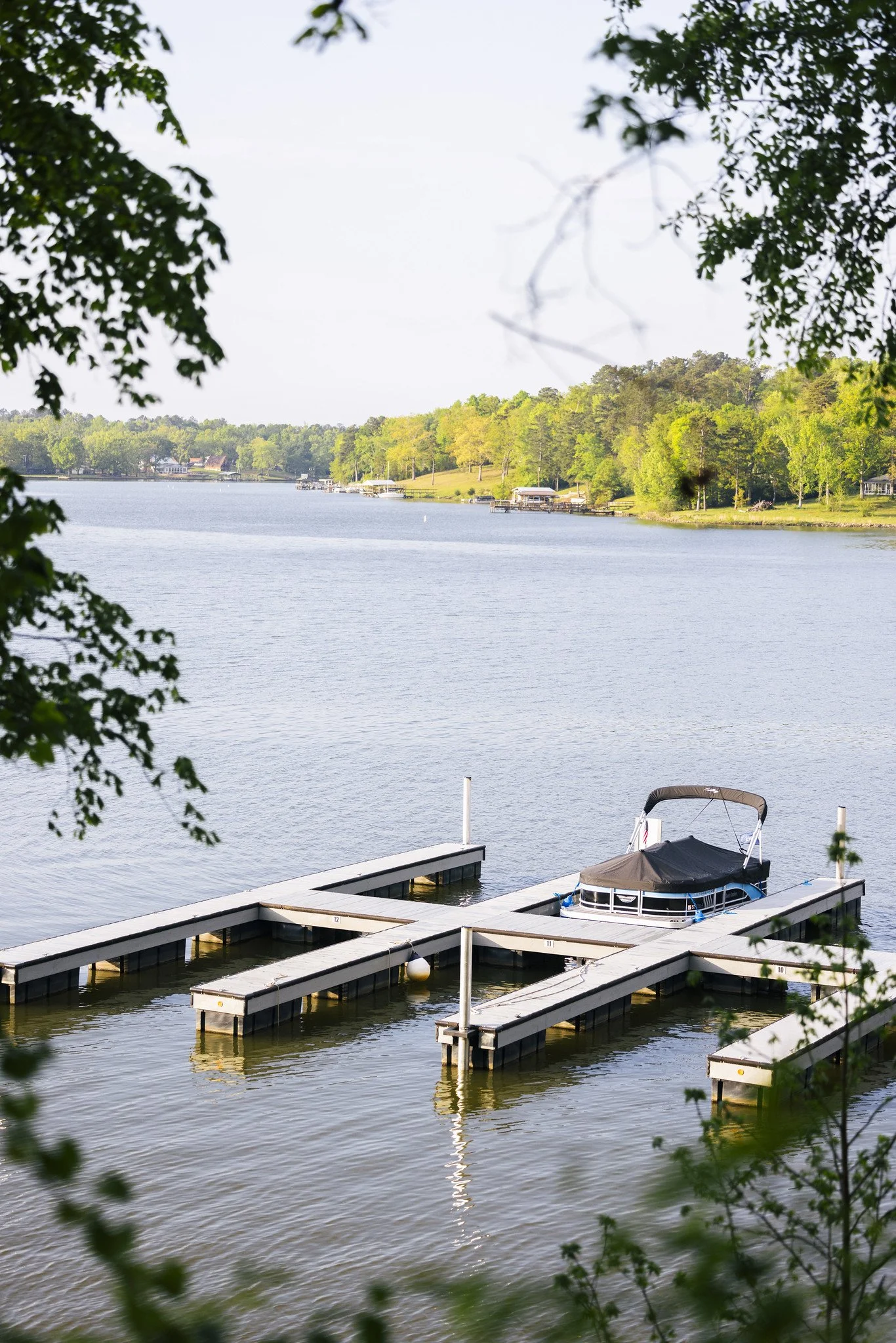 A marina with multiple boat docks on a river, one boat is docked, surrounded by trees and a distant shoreline with houses and more trees.