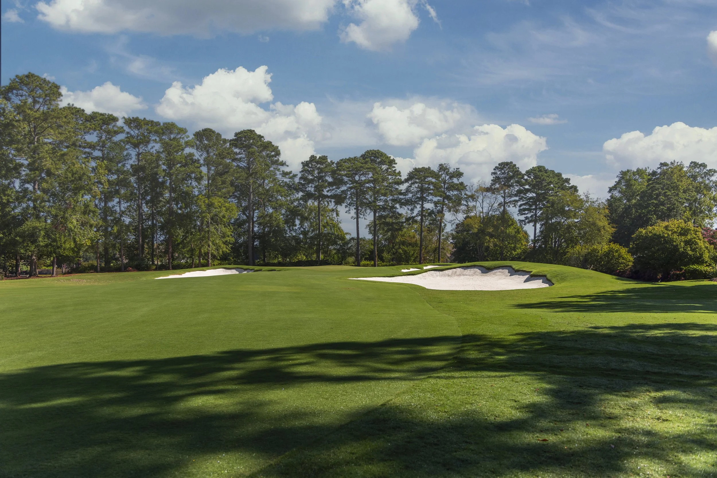 A golf course with green grass, sand bunkers, and trees in the background under a partly cloudy sky.