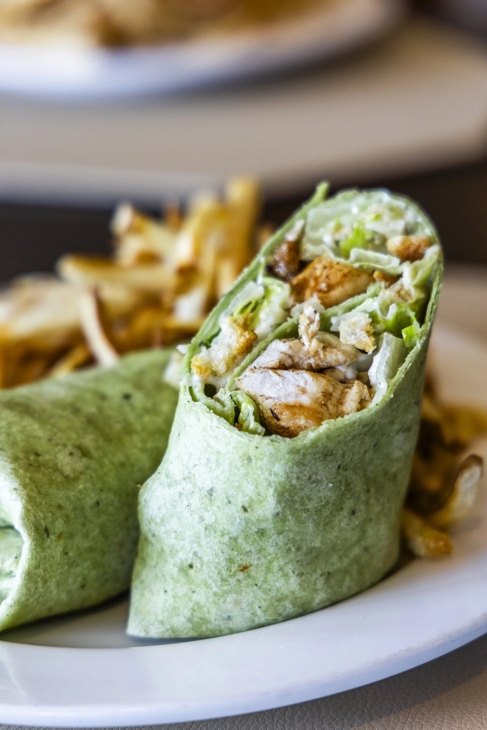 Close-up of a green wrap cut in half, revealing chicken, lettuce, and other ingredients inside, served on a white plate, with a side of fries in the background.