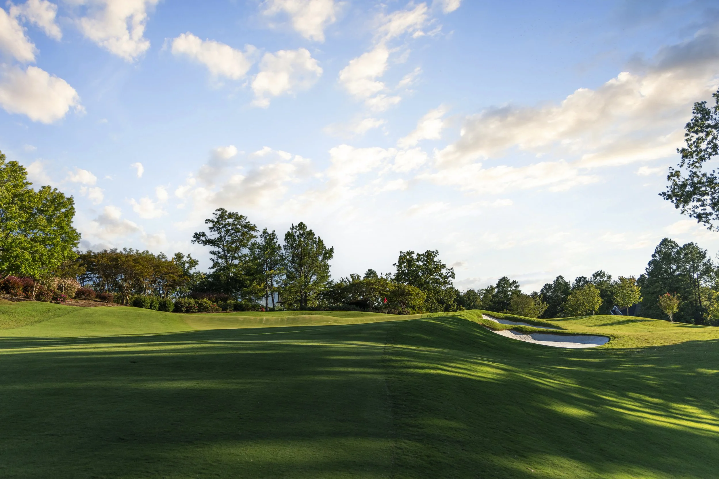 A golf course with a well-maintained green, sand bunkers, and trees under a partly cloudy sky.