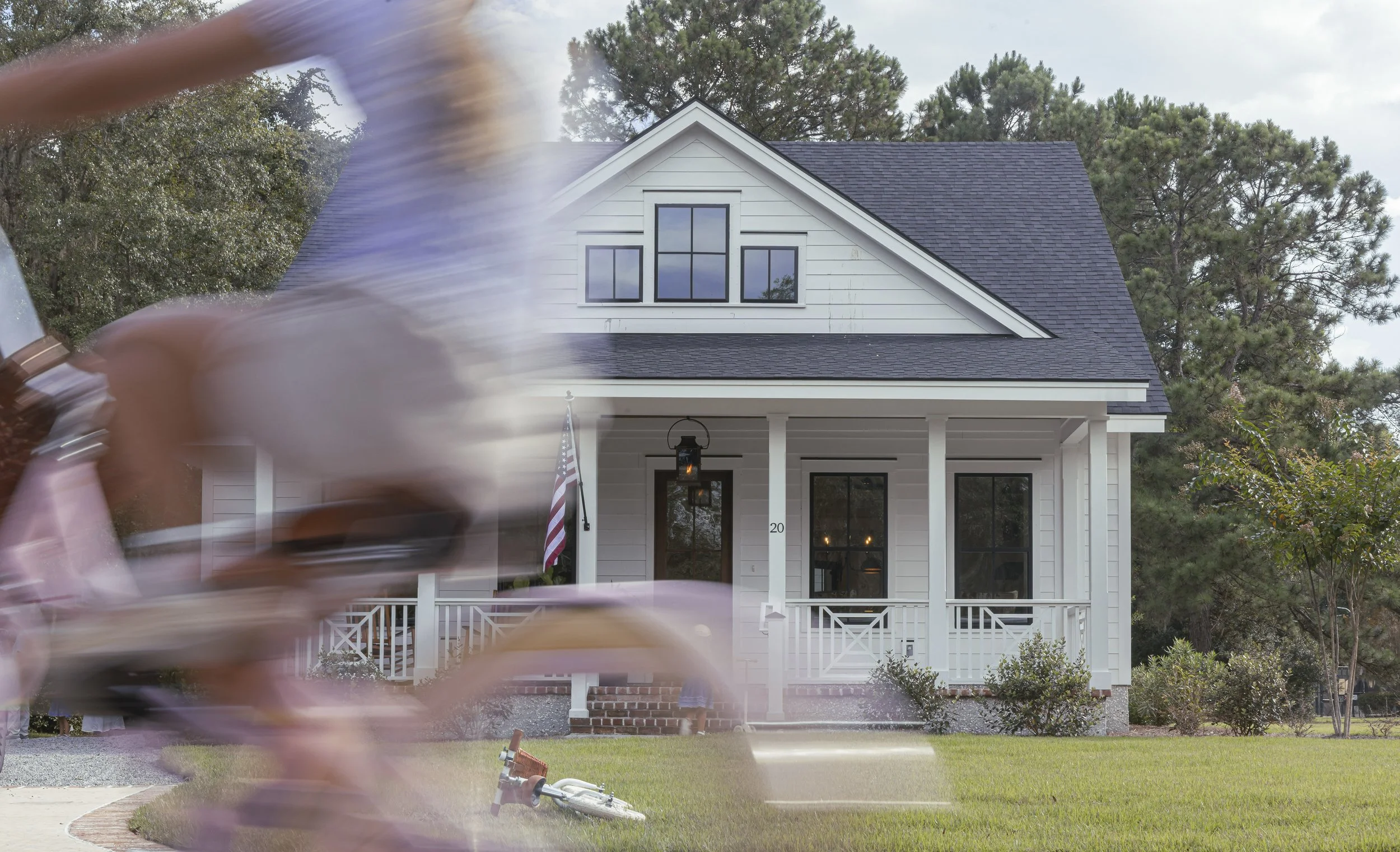 A blurred cyclist riding past a white house with a black roof in a suburban neighborhood.