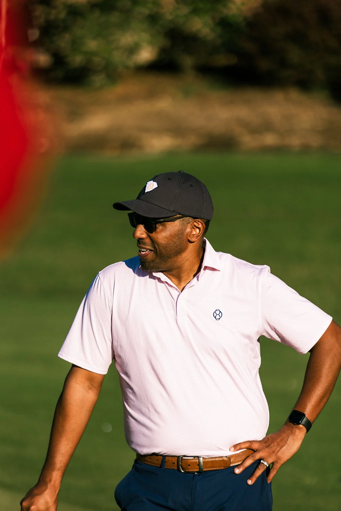 A man wearing sunglasses, a black baseball cap, a light pink golf shirt, and dark pants, standing outdoors on a grassy area.