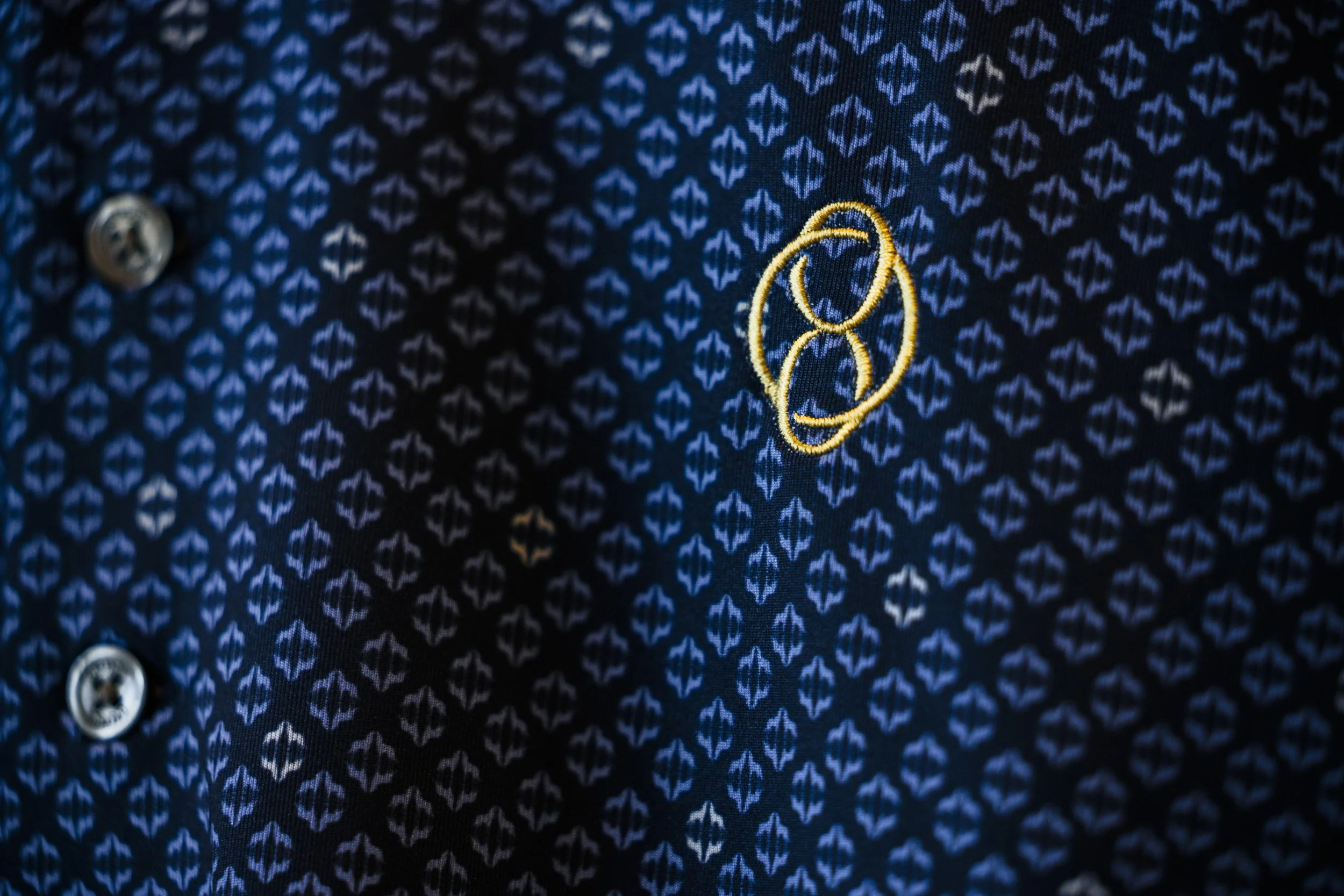 Close-up of a dark blue patterned fabric shirt with small white designs, a gray button on the left, and a yellow embroidered logo on the right.