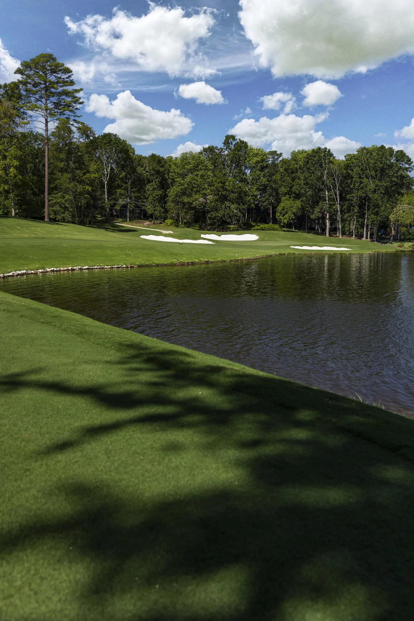 A golf course with water in the foreground, green grass, sand bunkers, trees, and a partly cloudy sky.