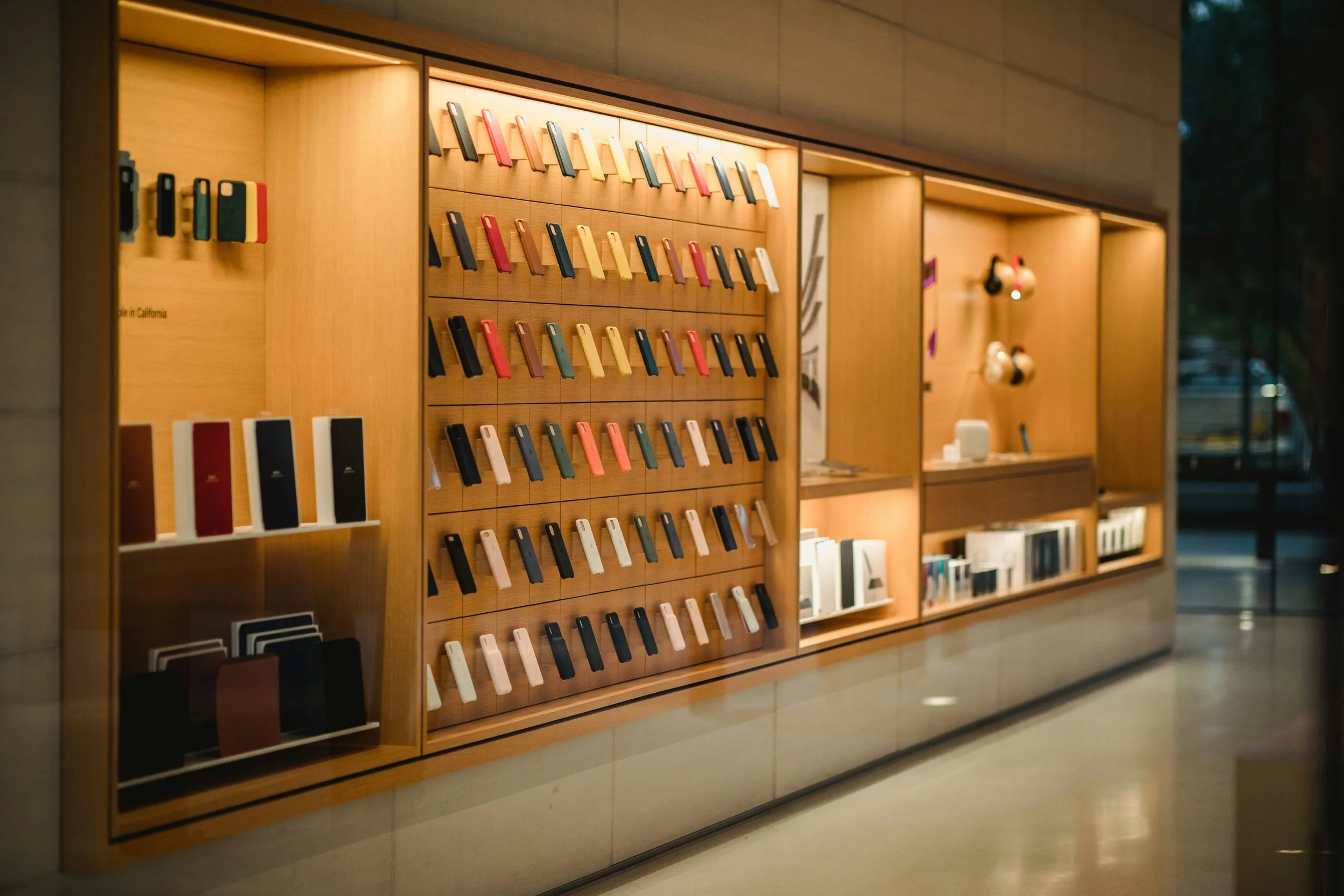Display of colorful phone cases arranged on wooden shelves in a retail store, with electronic accessories on the right side.
