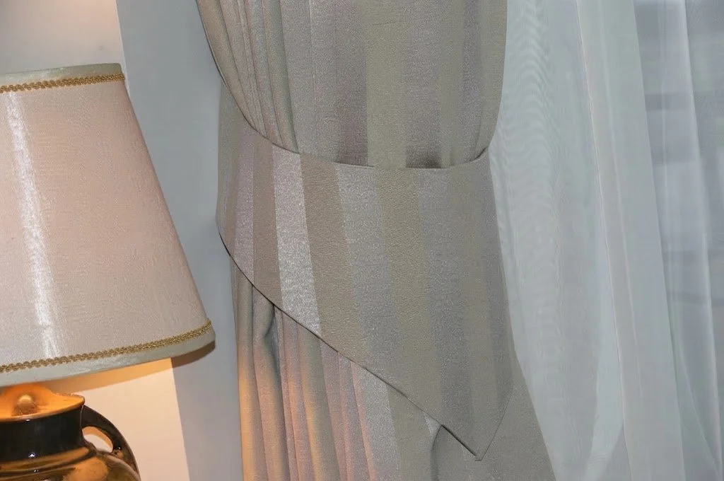 Custom drapery panel gathered with an elegant fabric tie-back, framing a classic bedside lamp and soft interior lighting by London Stitchery.