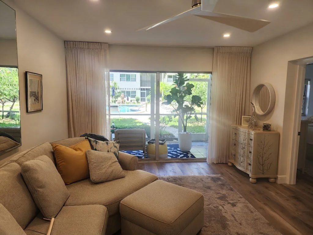 Custom sheer operational drapery panels framing a sun-filled living room with a sectional sofa and serene outdoor patio views, custom-crafted by London Stitchery.