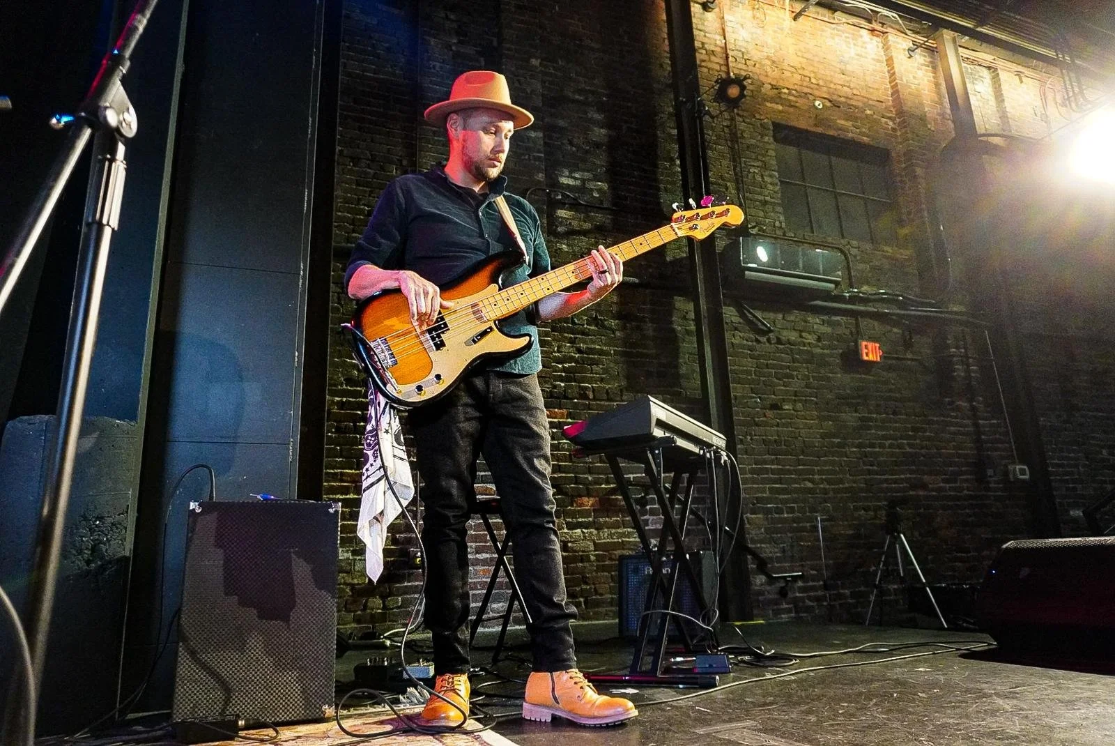 A man wearing a tan fedora and black shirt playing an electric bass guitar on a stage with a brick wall background, with music equipment around him.
