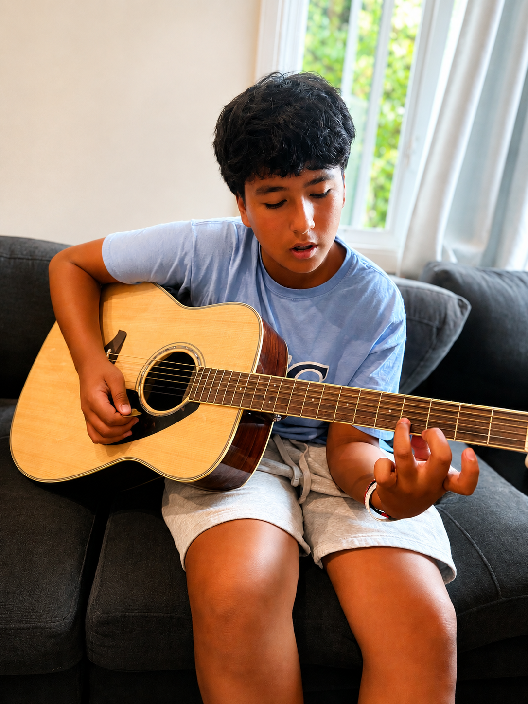 A young boy sitting on a black sofa, playing an acoustic guitar, with a window and greenery in the background.