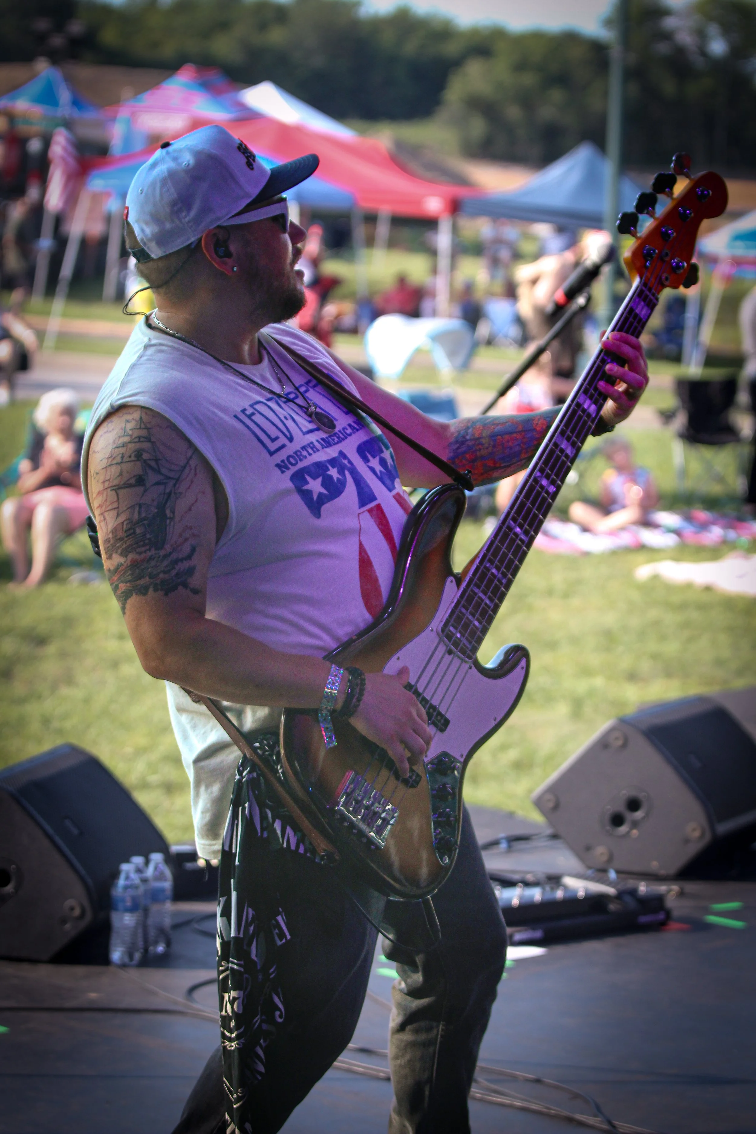 A man playing an electric bass on stage at an outdoor event with tents and people in the background.