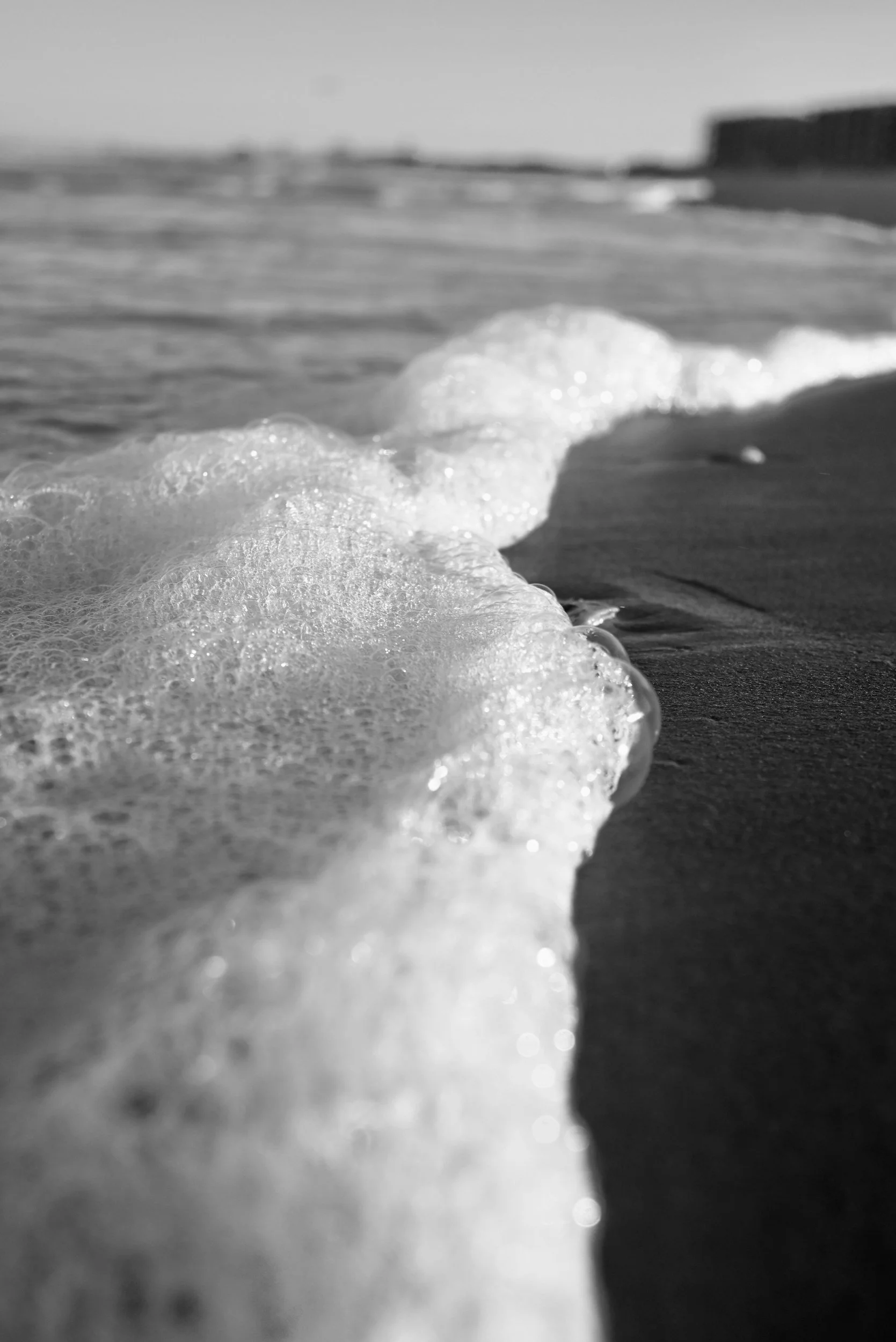 Black and white photo of ocean waves washing up on a sandy beach with foam, distant horizon, and cloudy sky.