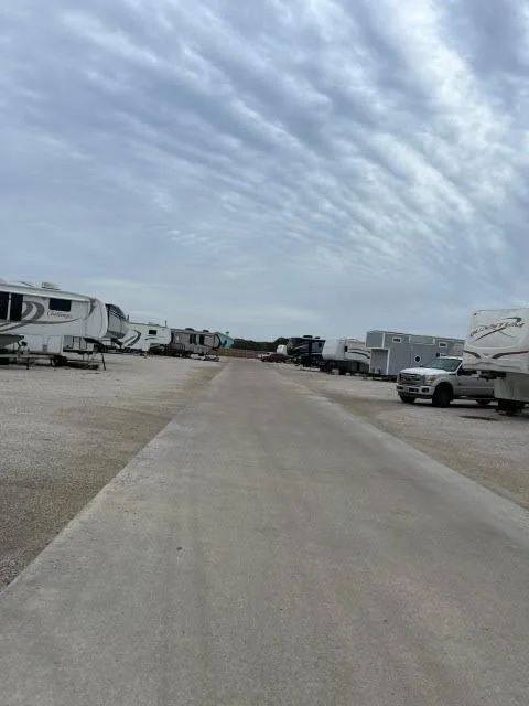 A gravel lot with parked RVs and trailers on both sides, and a gray truck driving down the center road under a cloudy sky.
