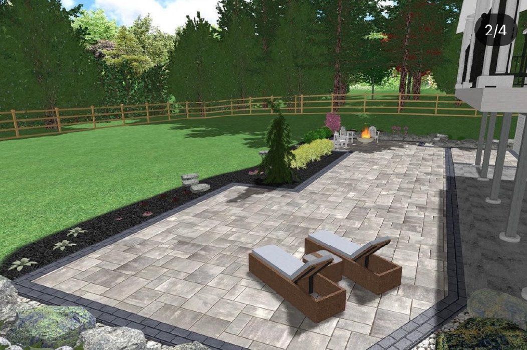 A backyard patio with beige pavers bordered with dark bricks, two lounge chairs with white cushions, a small fire pit with seating, and a lush green lawn surrounded by trees and a wooden fence.