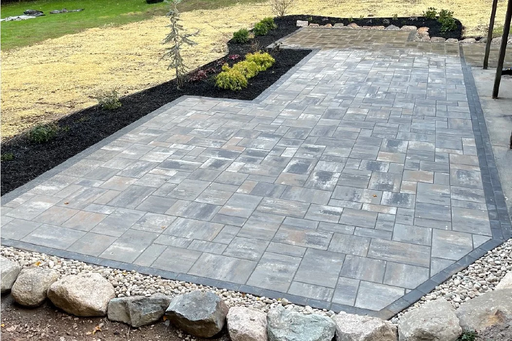A newly paved patio with gray stone tiles, surrounded by a border of small dark gray bricks, and a landscaped area with mulch, small plants, and a young tree in the background.